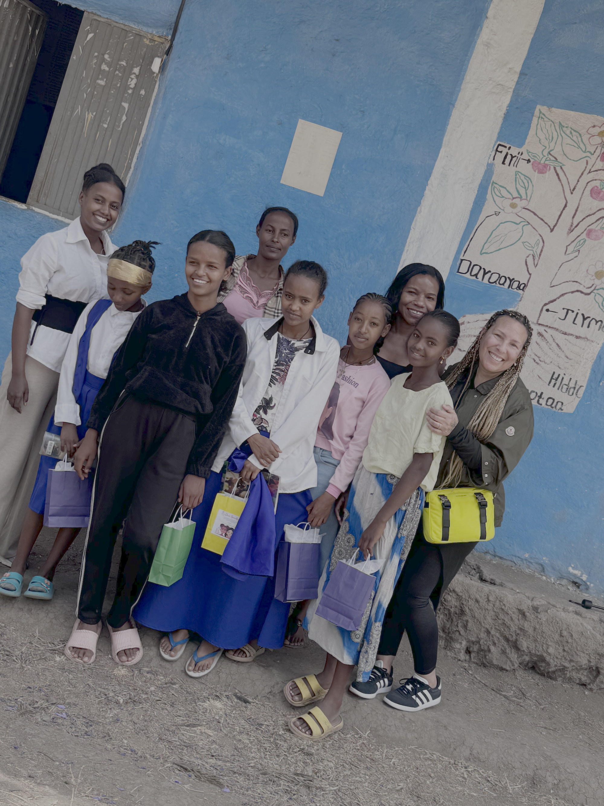Group of children and women standing in front of a bright blue painted wall, smiling and holding colorful shopping bags, with some wearing casual clothing and sandals.