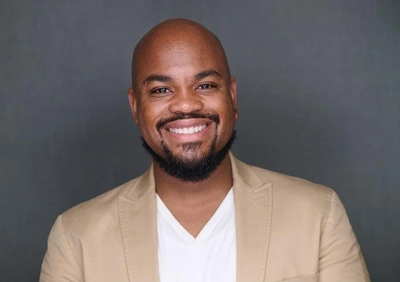 A man with a shaved head and beard smiling, wearing a tan blazer over a white shirt, against a gray background.