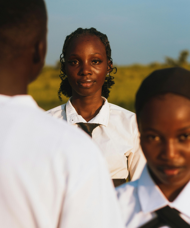 A young woman with curly hair and a white shirt stands outdoors, looking directly at the camera. Two other individuals are in the foreground, partially visible, wearing white shirts. The background features a natural landscape with greenery and a clear sky.