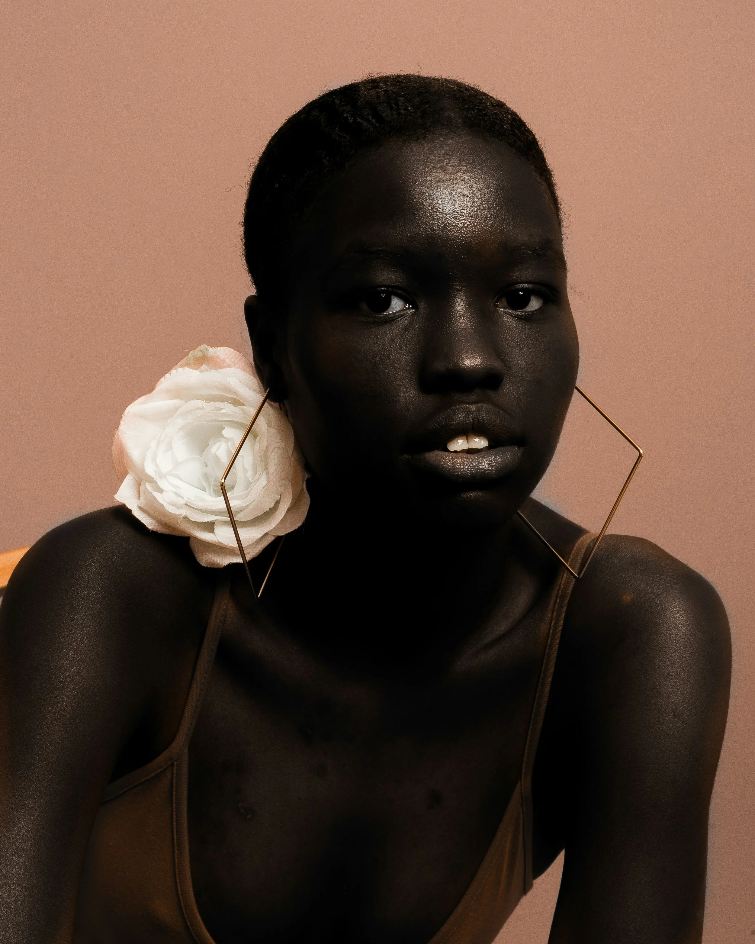 A close-up portrait of a black woman with short curly hair, wearing a black sleeveless top and a large white flower earring with an abstract gold frame, against a plain beige background.