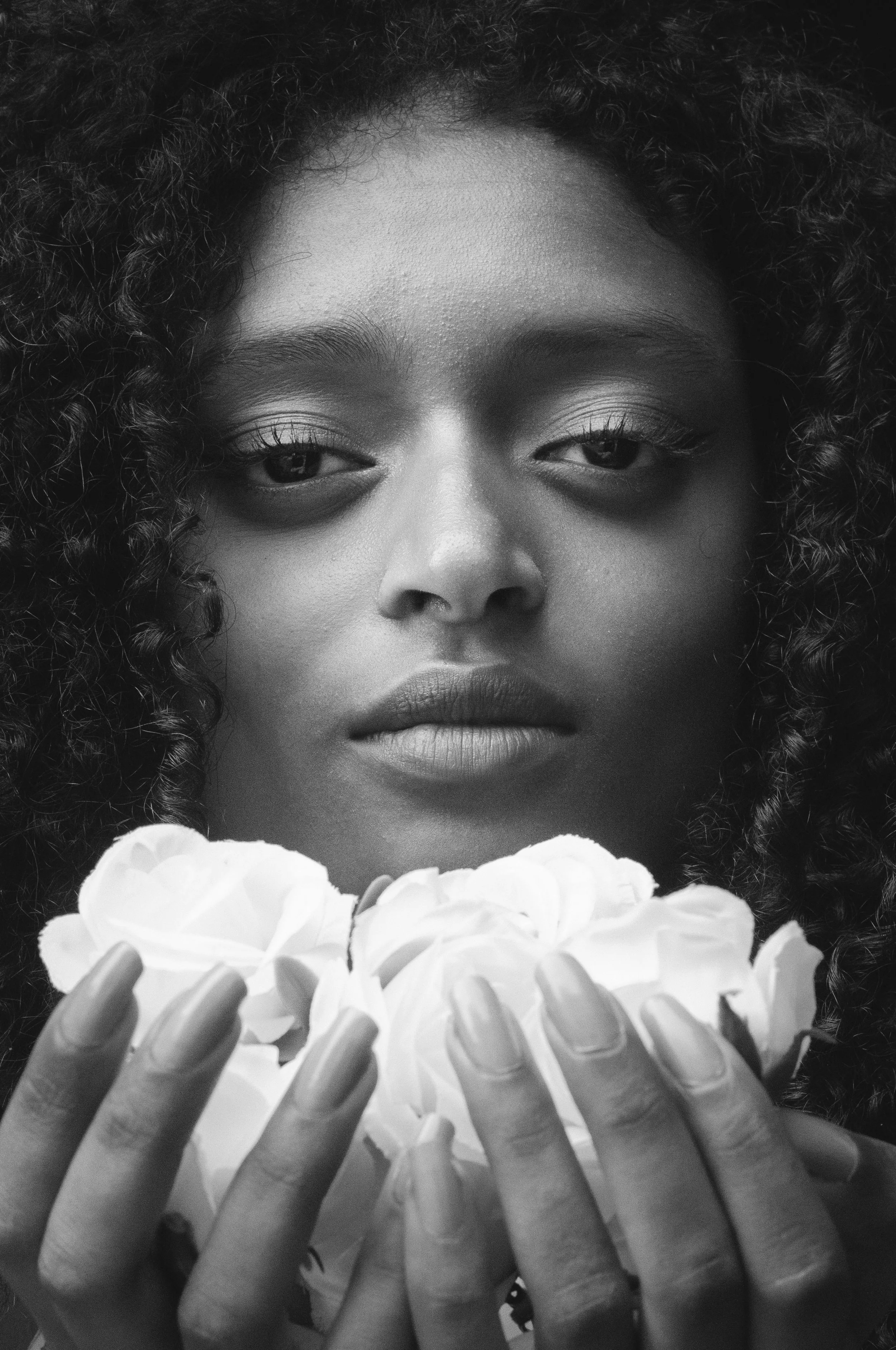A close-up black-and-white photo of a woman with curly hair holding a bunch of flowers with both hands, partially covering her face.