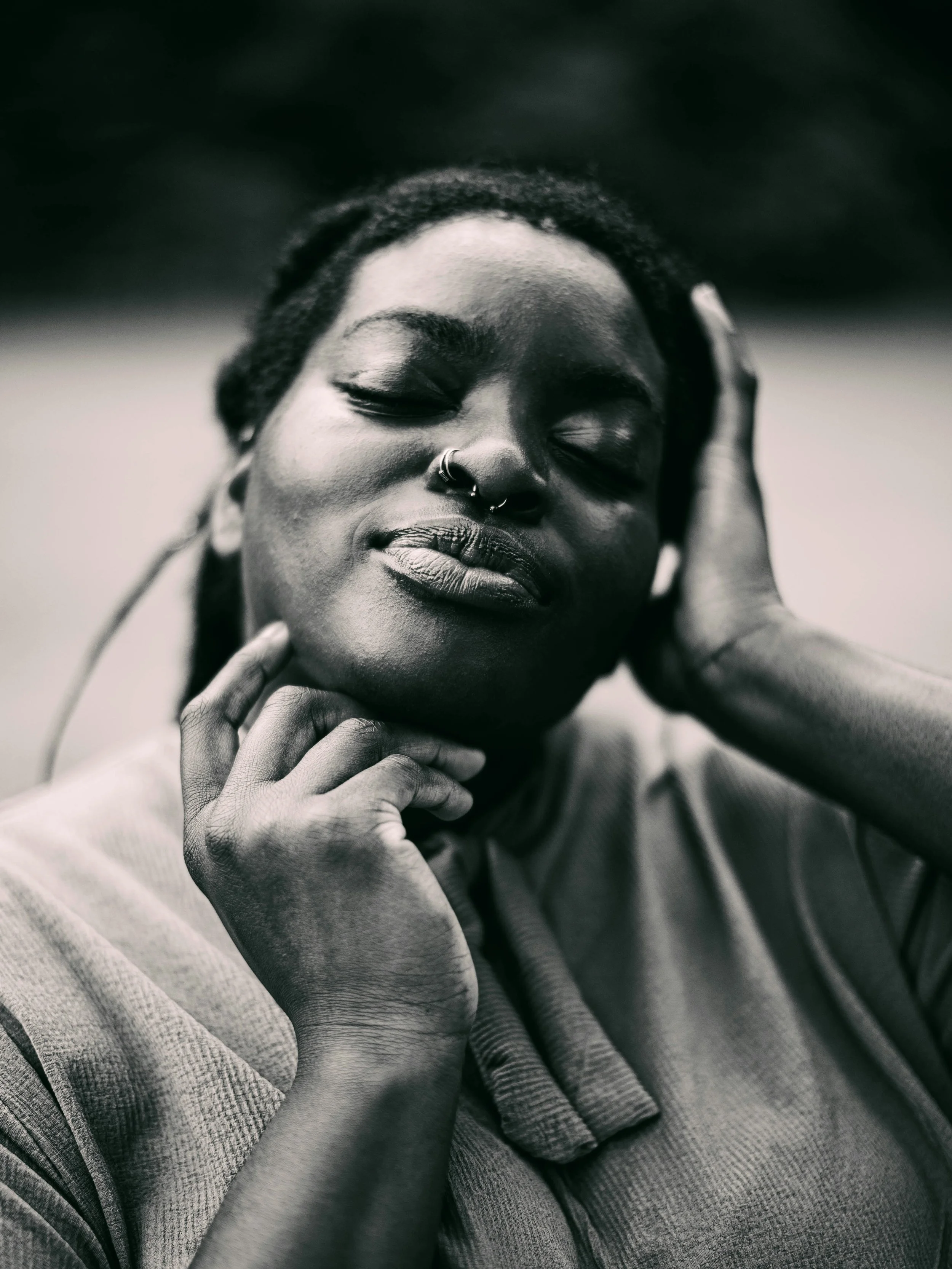 Close-up black and white photo of a person with closed eyes, relaxed expression, nose rings, touching their face and head, wearing a long-sleeve shirt.