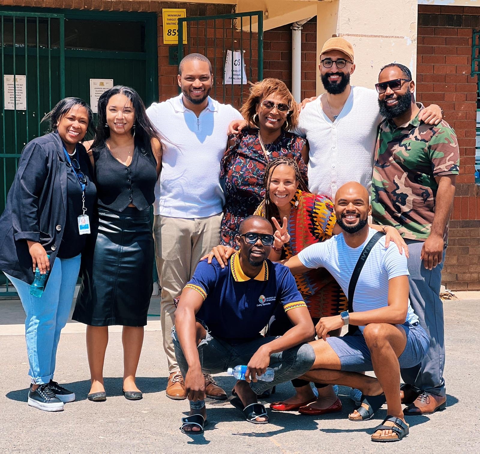 A diverse group of eleven people posing outdoors in front of a brick building, smiling and making various gestures. Some are standing, while others are sitting or squatting. They appear to be friends or colleagues enjoying a sunny day.