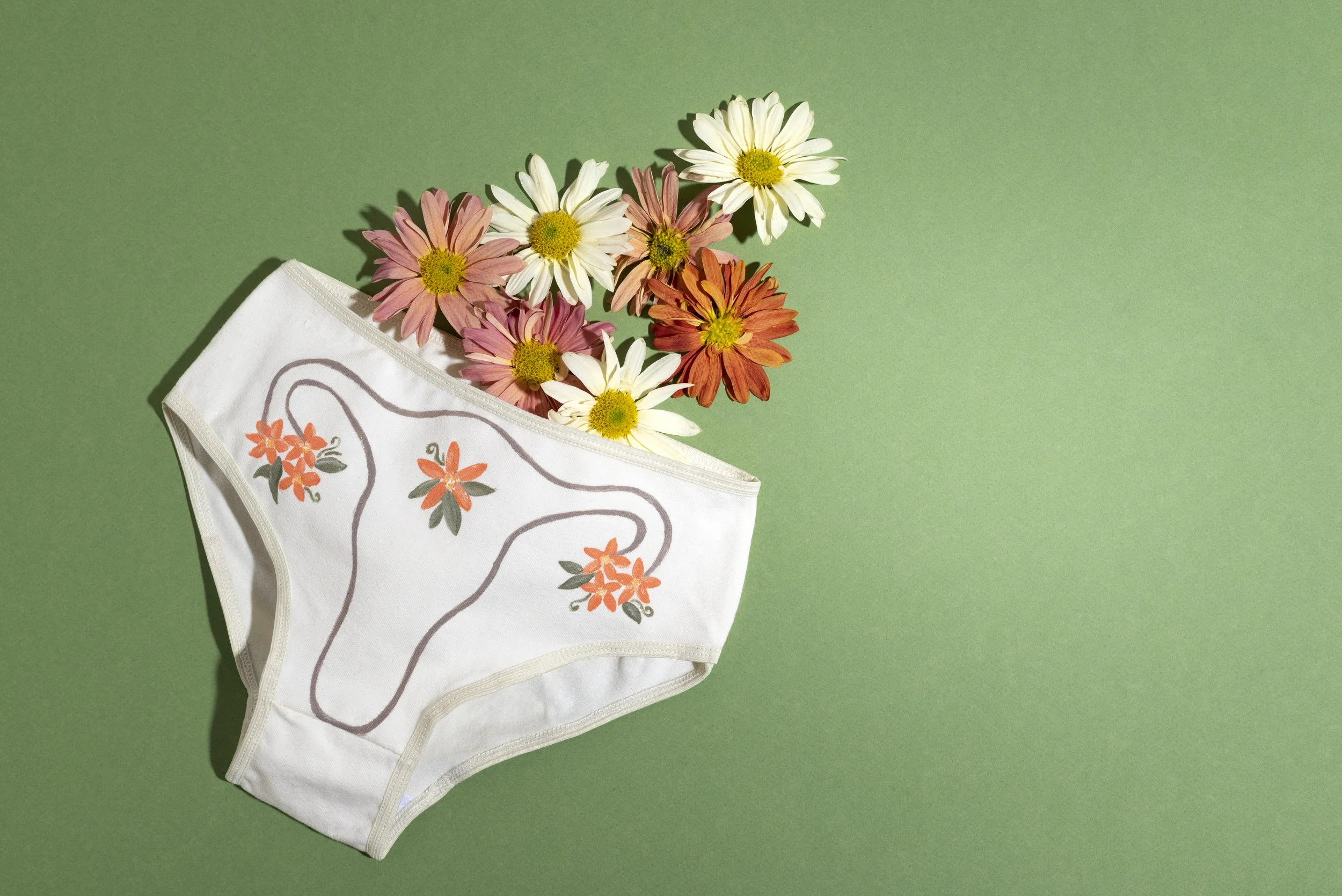 A paper face mask with flower designs hanging from a bouquet of daisies against a light green background.
