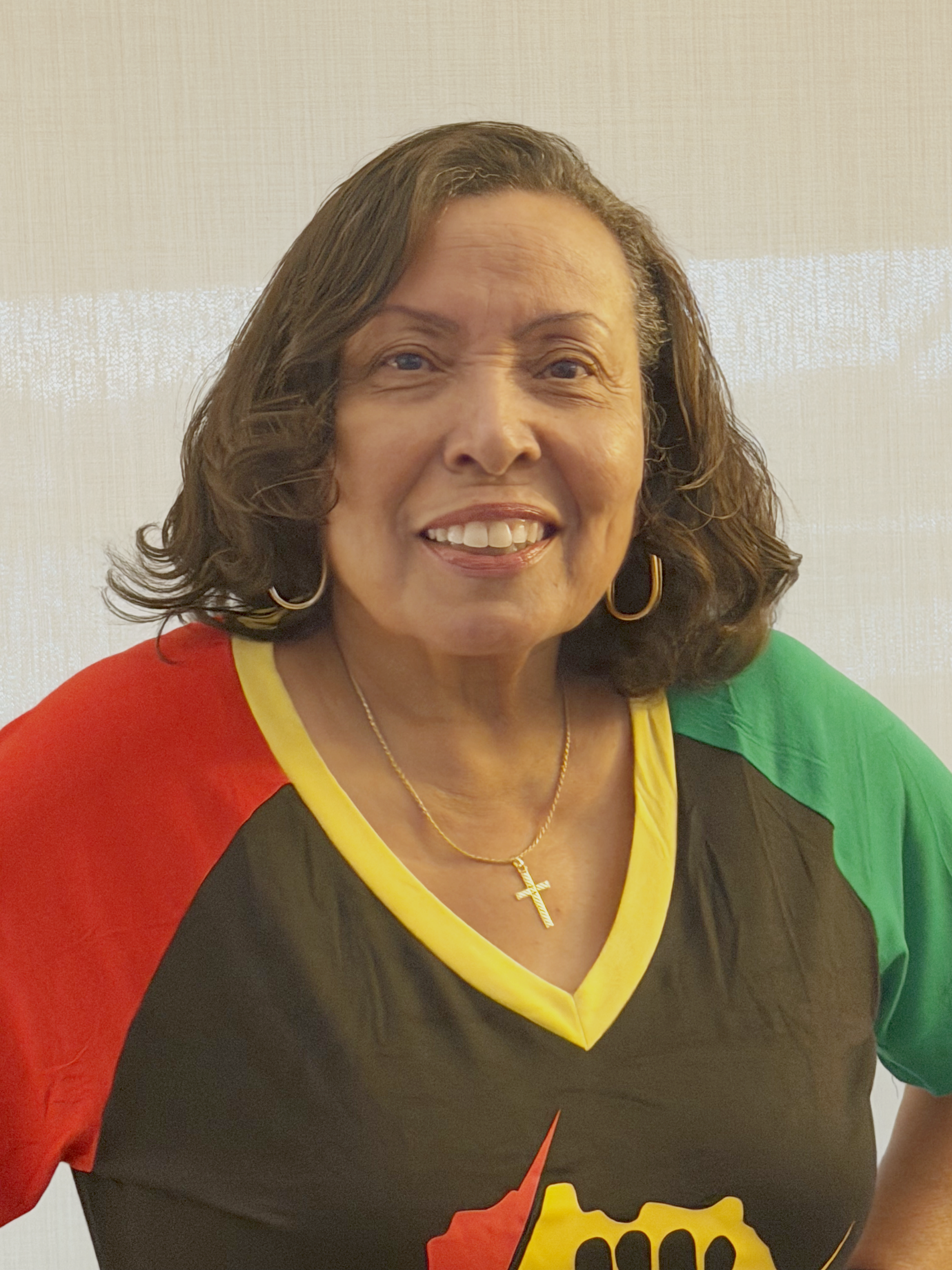 Close-up of a woman with curly dark hair, smiling, wearing a red floral shirt, gold hoop earrings, and a gold cross necklace, with a light green background.