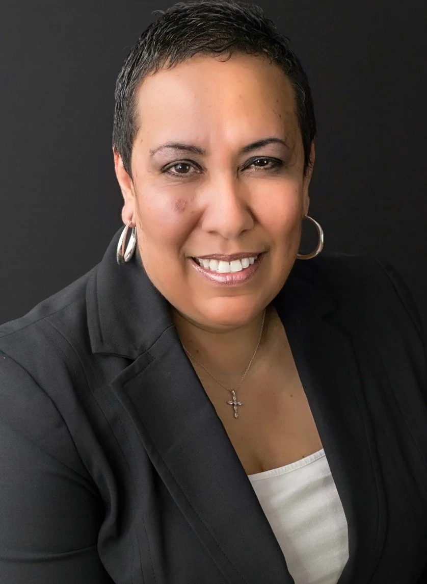 Close-up portrait of a woman with short dark hair, wearing a black blazer, white top, hoop earrings, and a cross necklace, smiling against a black background.