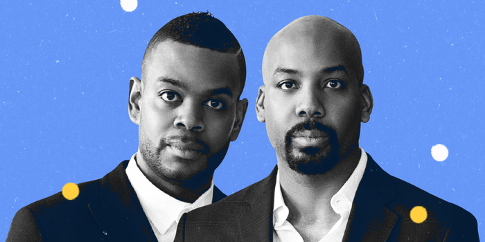 Two men wearing suits against a blue background with white and yellow dots.