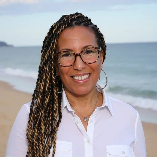 A woman with long braided hair, wearing glasses and a white shirt, smiling on a beach with the ocean and sky in the background.