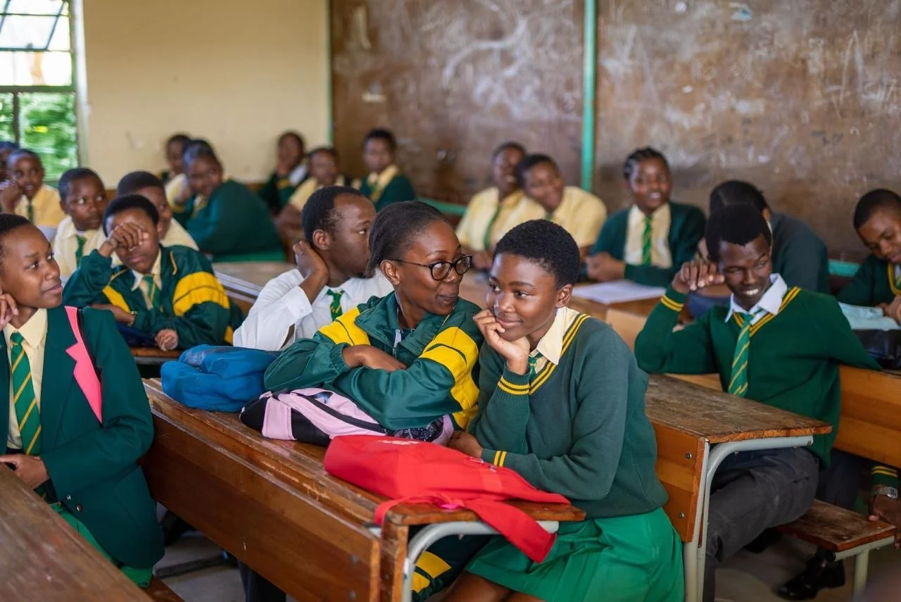 A classroom full of students wearing green and yellow uniforms, sitting at wooden desks, with some smiling and chatting with each other.