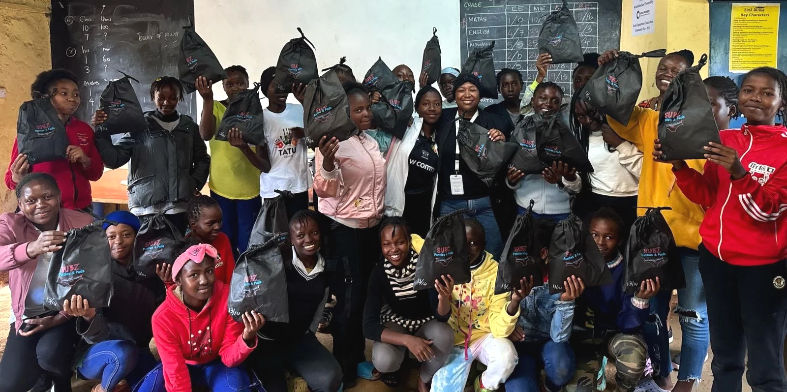 Group of children and two adults in a classroom holding black bags with a logo. The children appear happy, and the classroom has posters and a blackboard with writing.