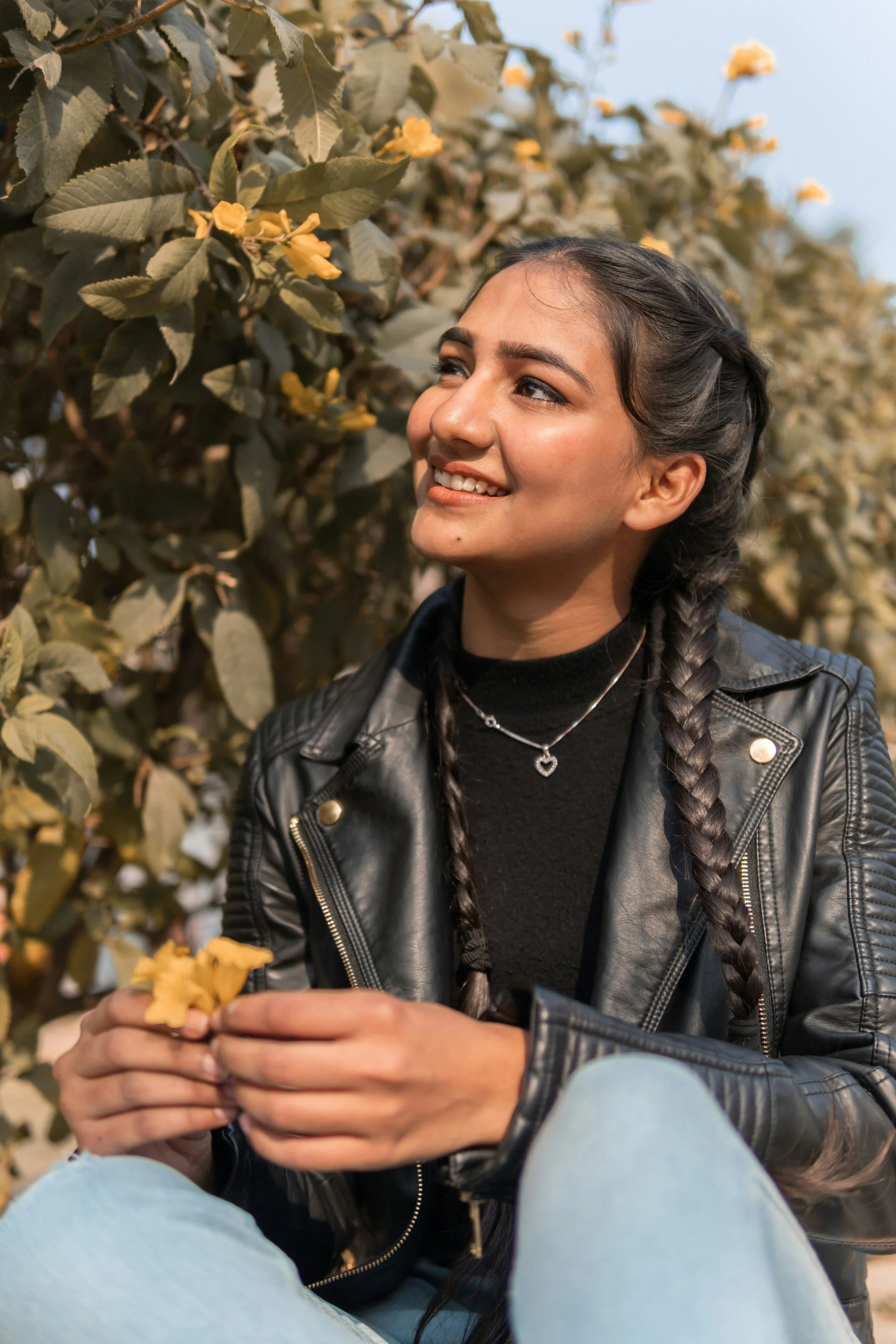 A smiling young woman with braided hair, wearing a black leather jacket, sitting outdoors among green shrubbery with yellow flowers.