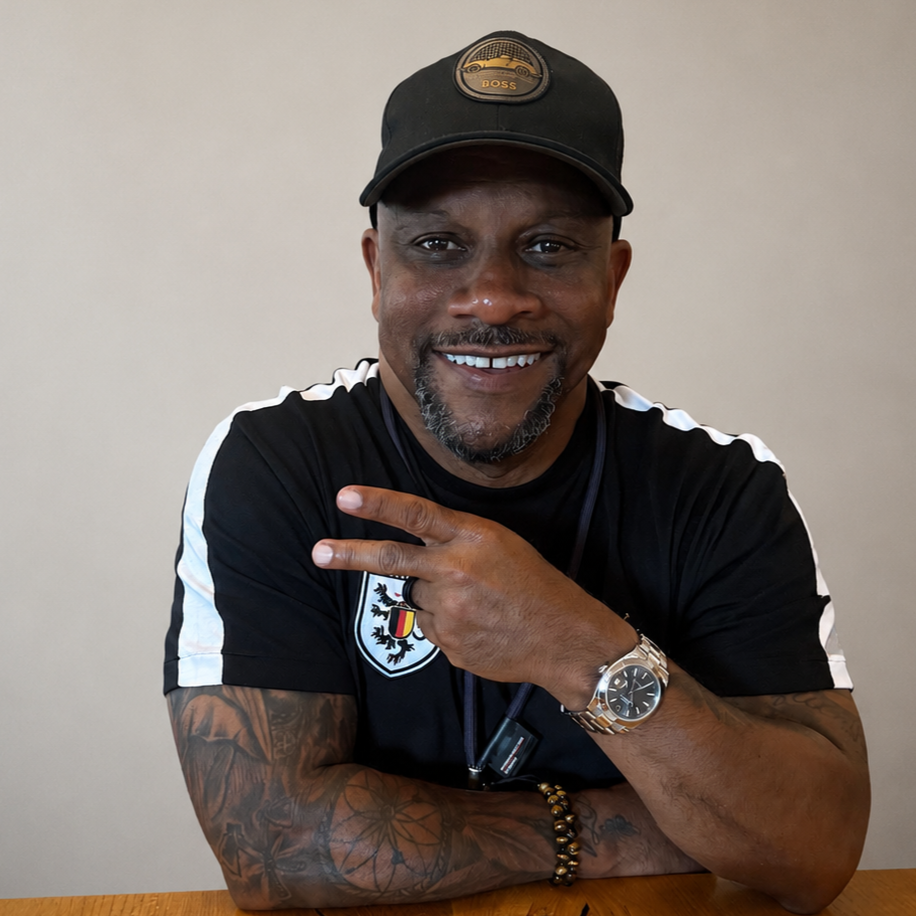 A smiling man wearing a black cap with a logo, a black sports shirt with a crest, a silver watch, and bracelets, making a peace sign with his fingers.