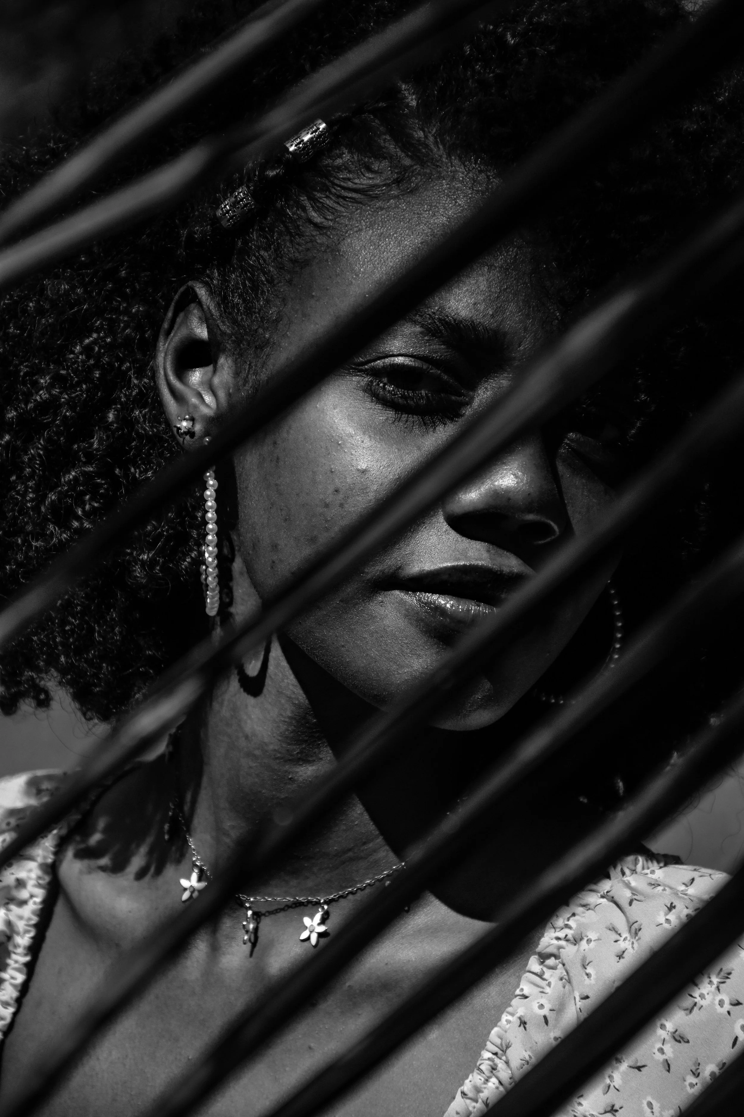 A black and white photograph of a woman with curly hair, wearing earrings and a necklace, looking through bars.