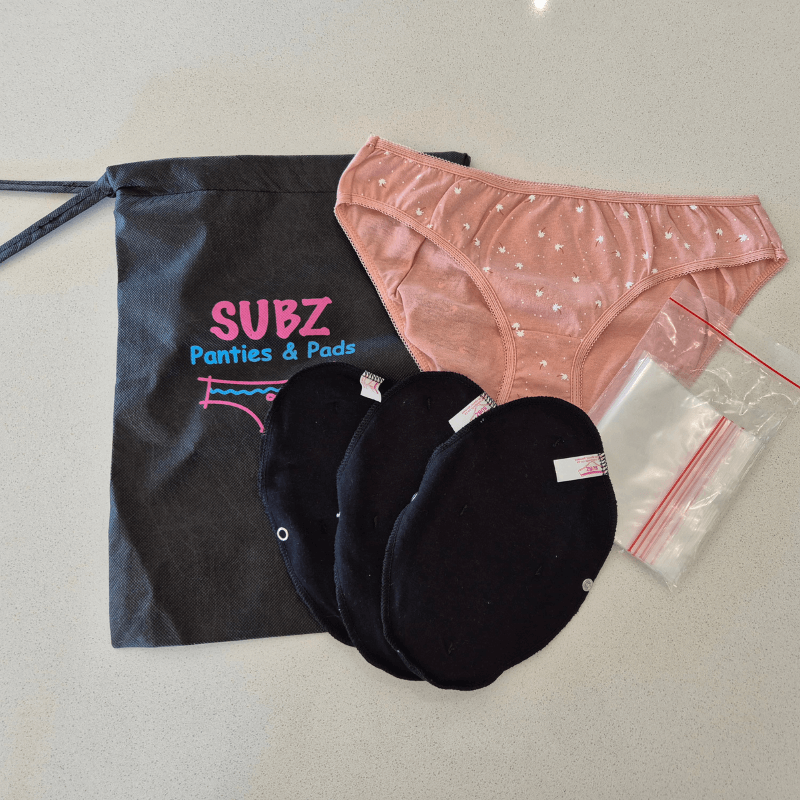Pink panties with white stars, three black panty liners, a black storage bag labeled 'SUBZ Panties & Pads', and a small plastic resealable bag on a gray surface.
