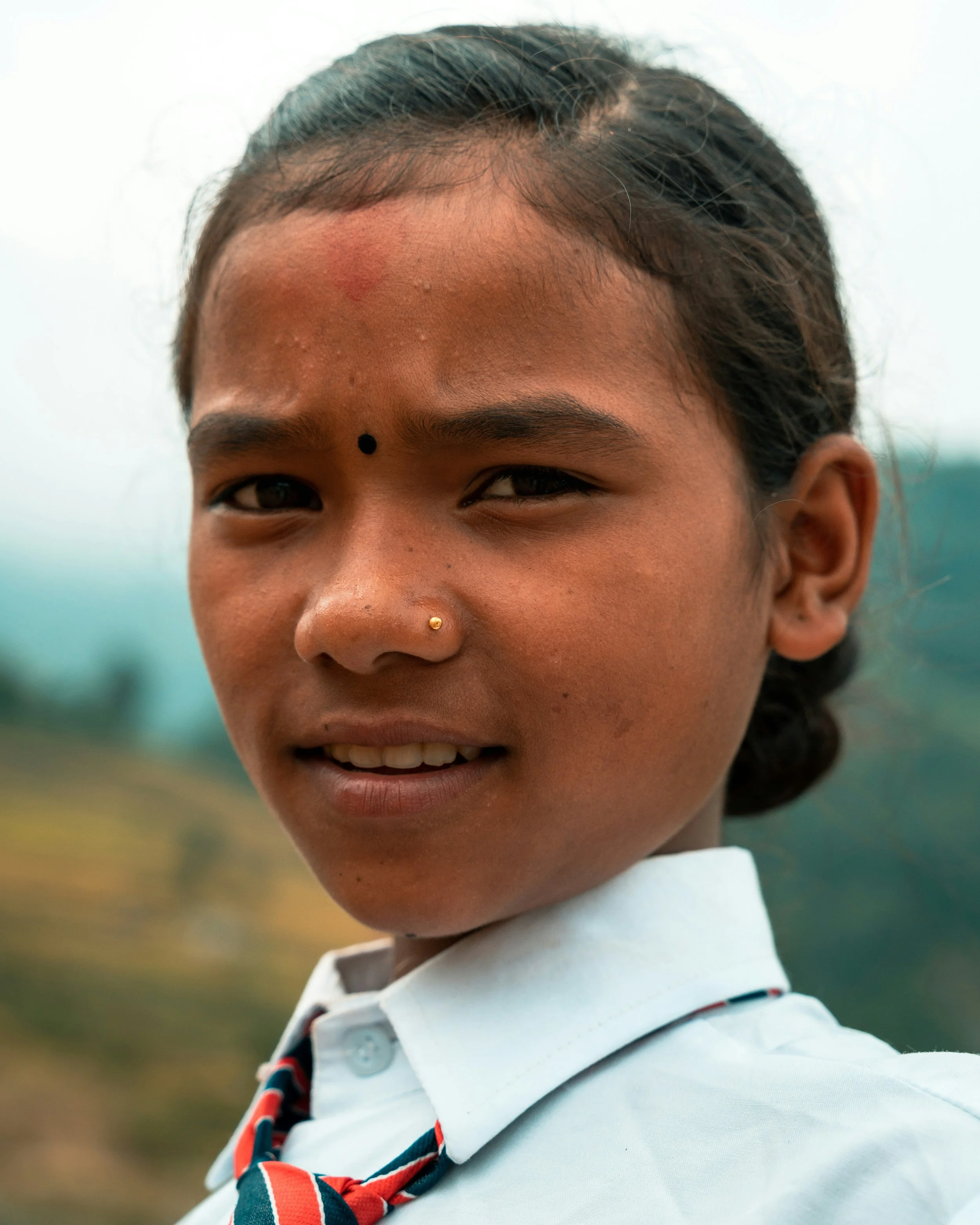 Close-up of a young girl wearing a white shirt, striped tie, and a nose piercing, smiling outdoors with a blurred green background.