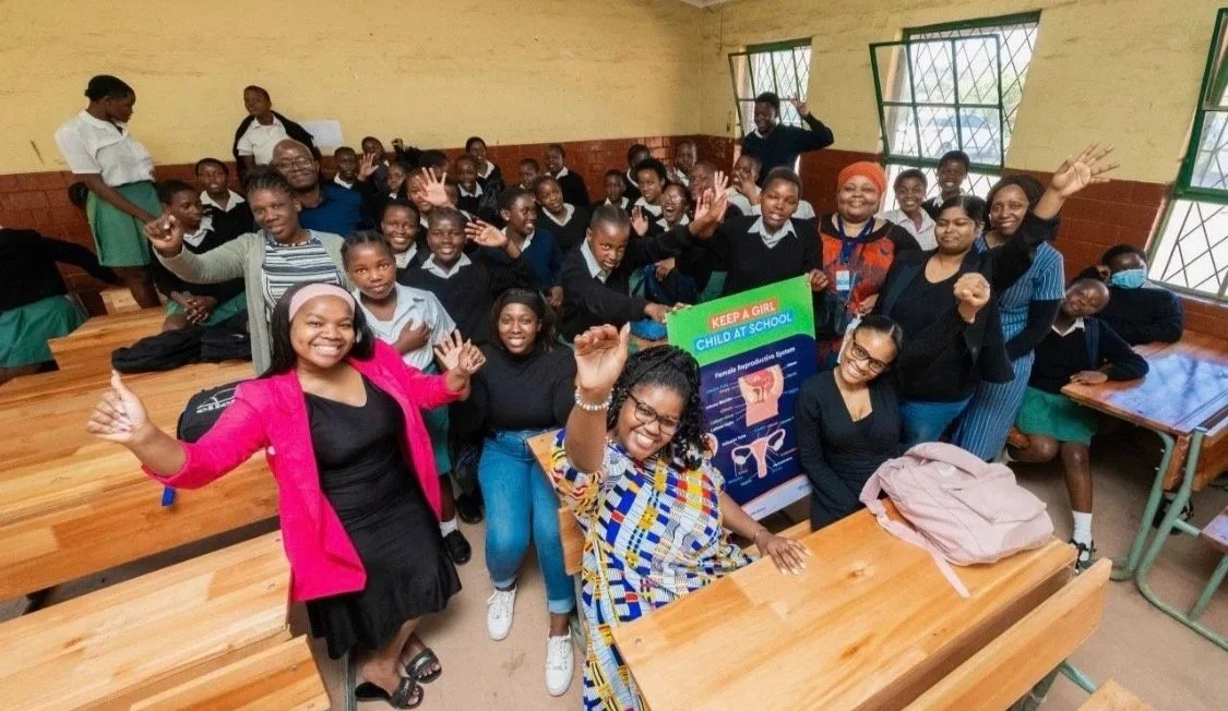 A classroom filled with students and teachers smiling and raising their hands, with a poster about female reproductive health.
