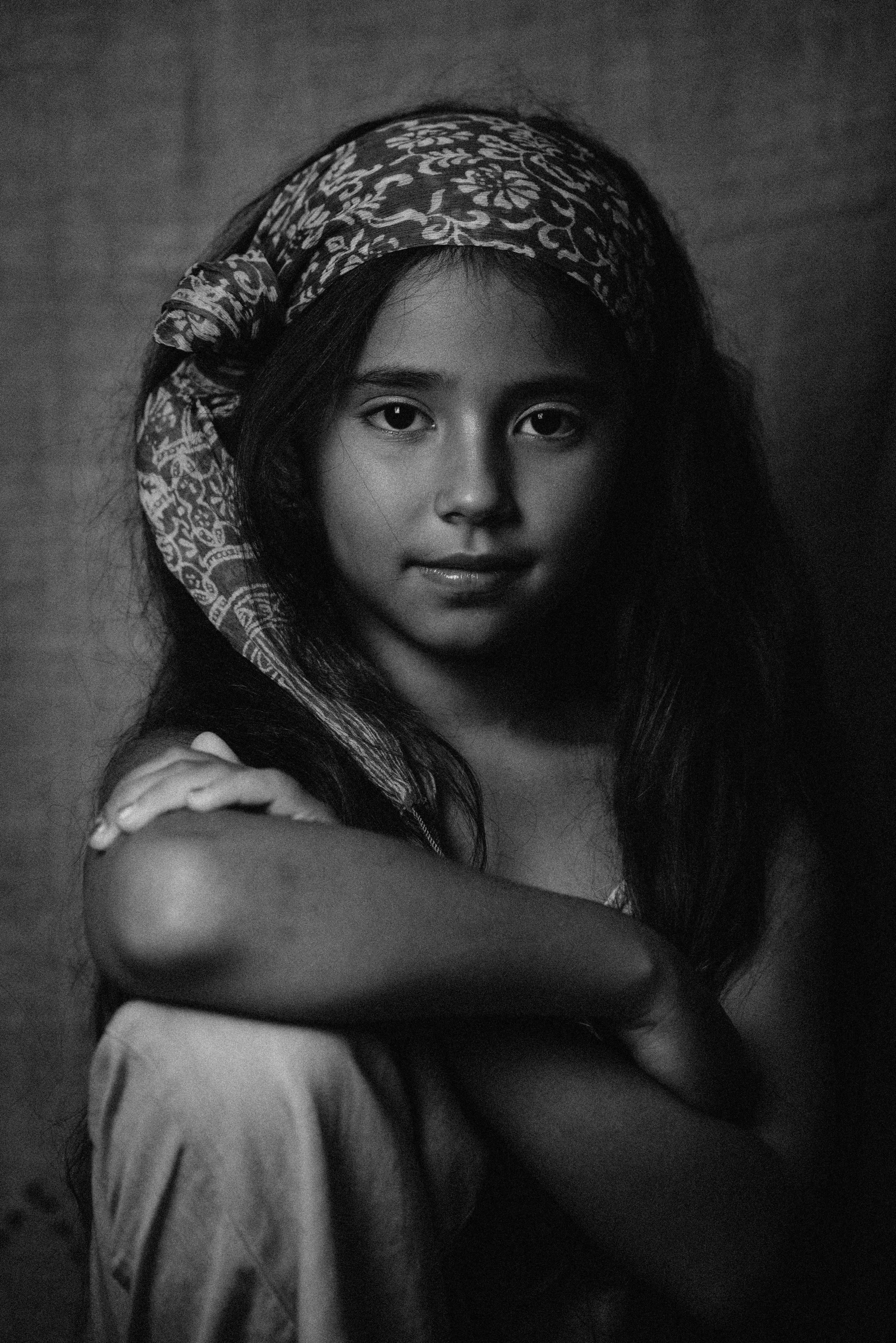 Black and white photograph of a young woman with long hair, wearing a patterned headscarf, looking directly at the camera with a calm expression, her arm crossed in front of her.