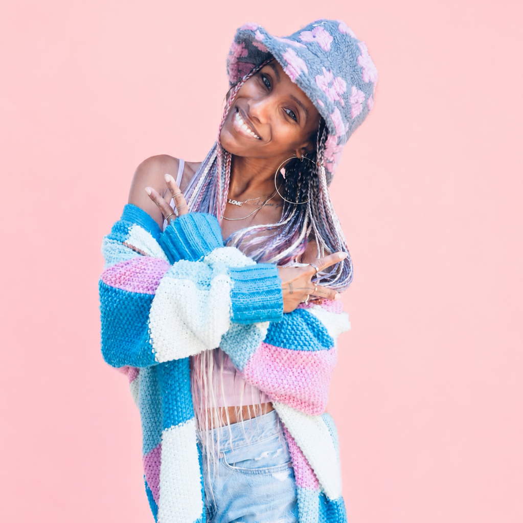 A young woman smiling and posing with crossed arms in front of a pink background. She is wearing a colorful knit sweater, distressed jeans, and a fuzzy hat with pink and purple patterns. She has long braided hair with colorful extensions, large hoop earrings, and several necklaces.