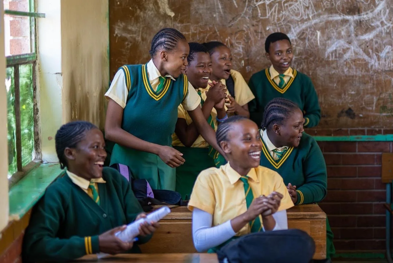 Group of students wearing yellow shirts and green vests or sweaters, sitting and standing in a classroom, smiling and laughing.