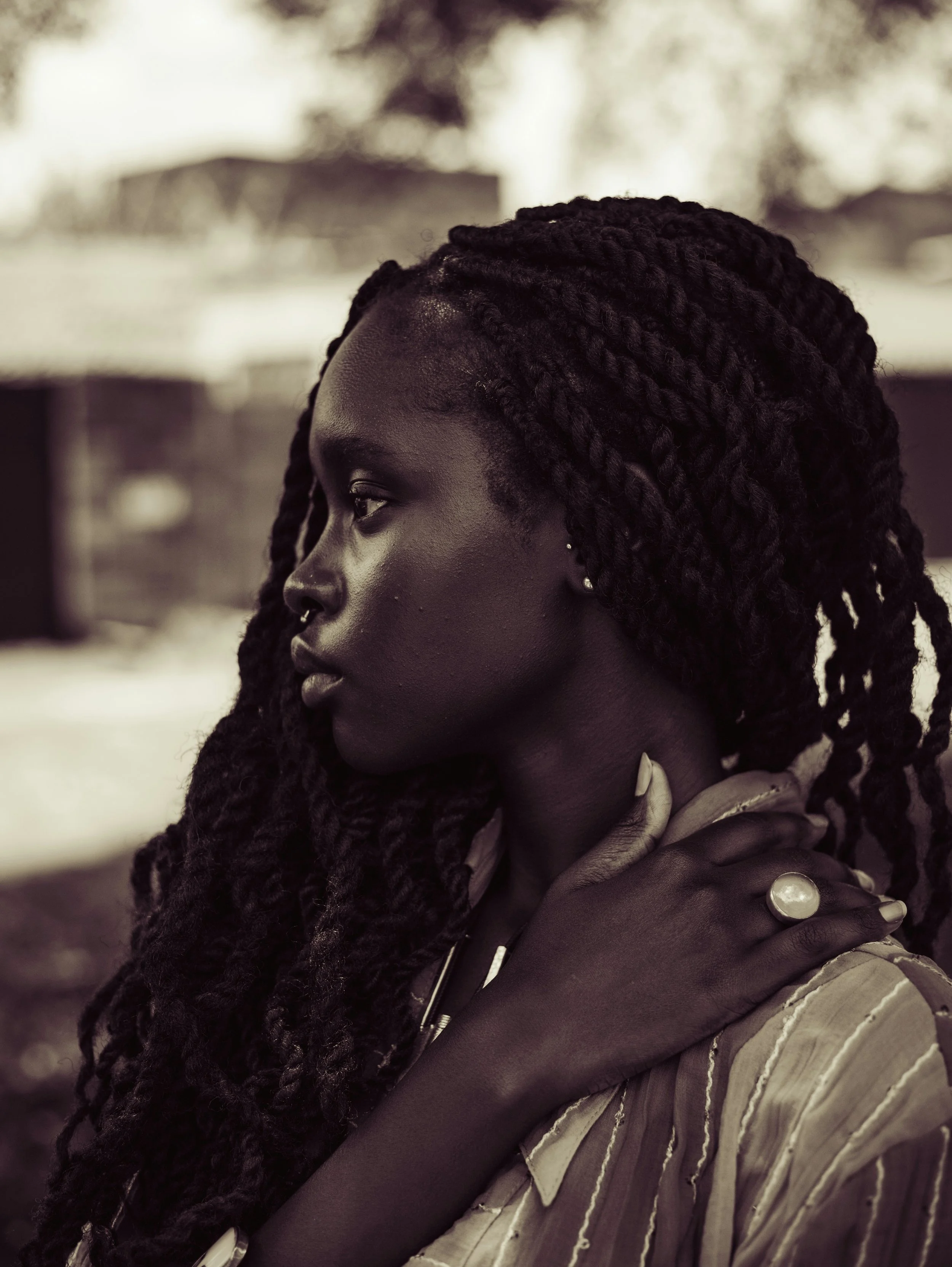 A woman with dark skin and long, braided hair, wearing a striped shirt, is outdoors with a blurred background.