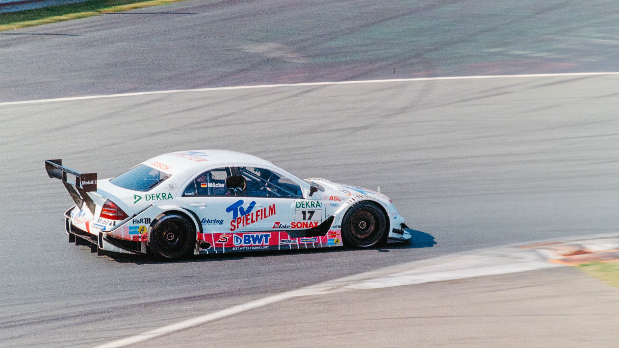 A white race car with various sponsor logos, including TV Spielfilm and BWT, speeds on a racetrack with motion blur in the background.