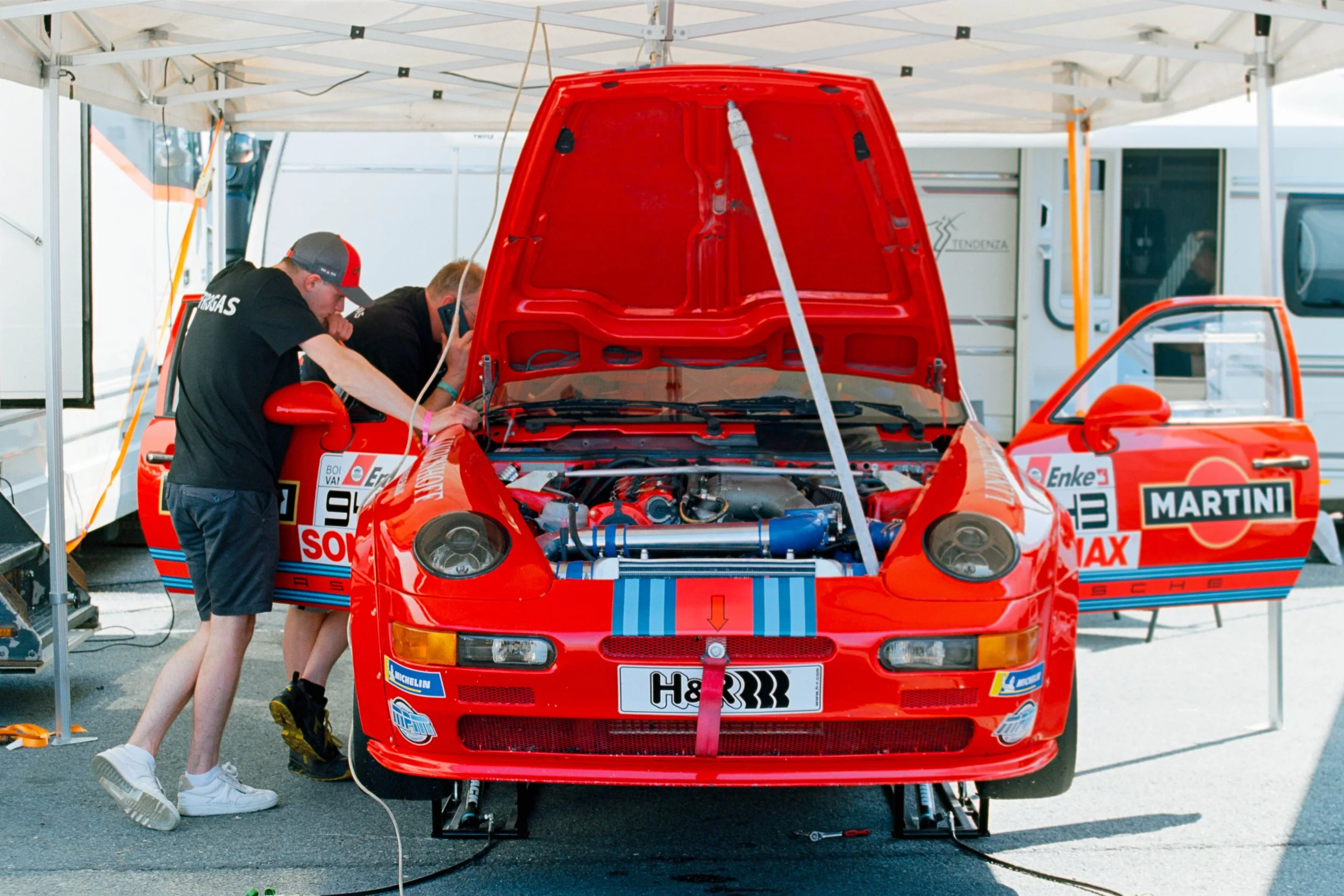 Two mechanics working on a red race car with its hood open under a tent at a racetrack.