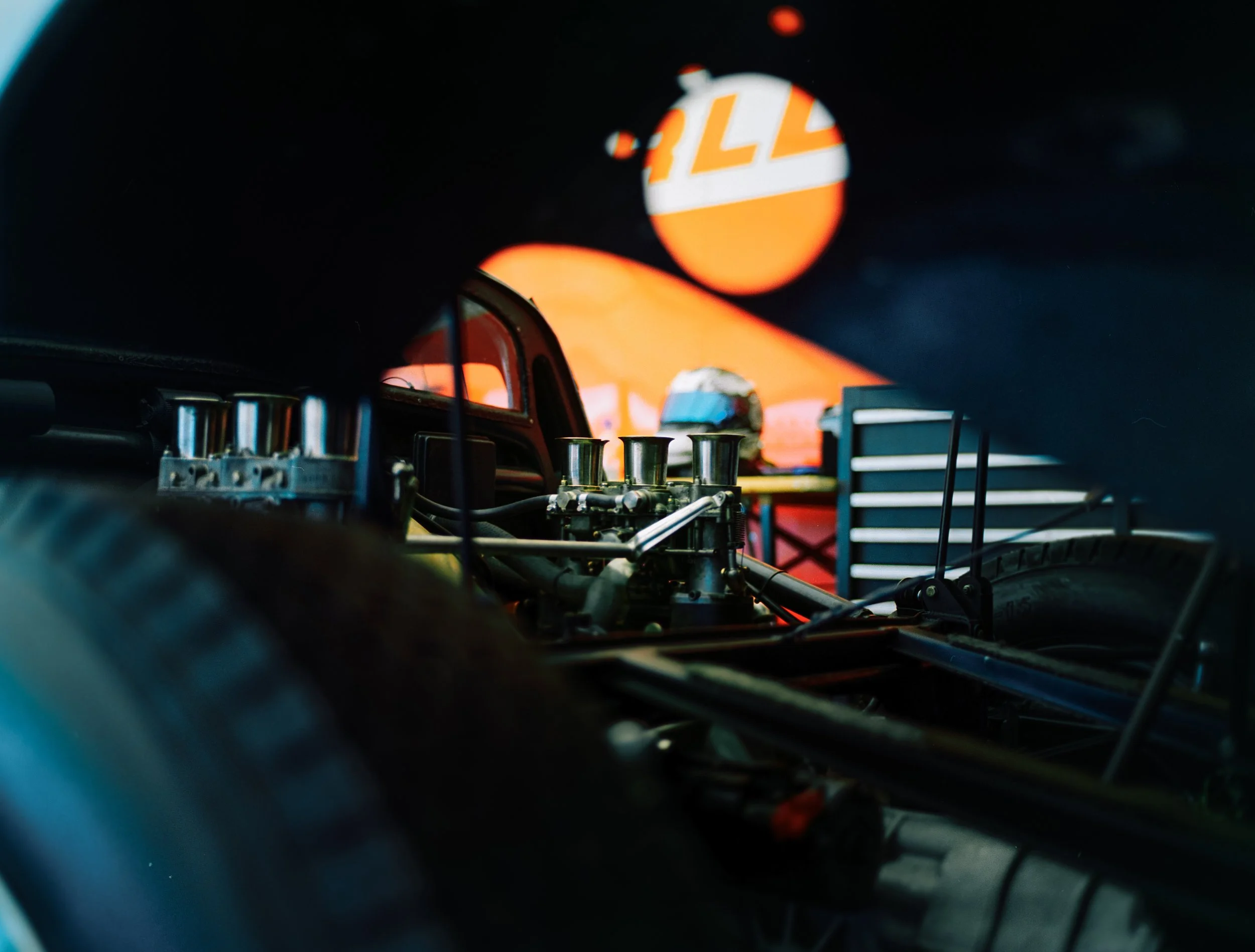 Close-up of a vehicle dashboard with orange gauges and controls, and a blurred view of a race car in the background.