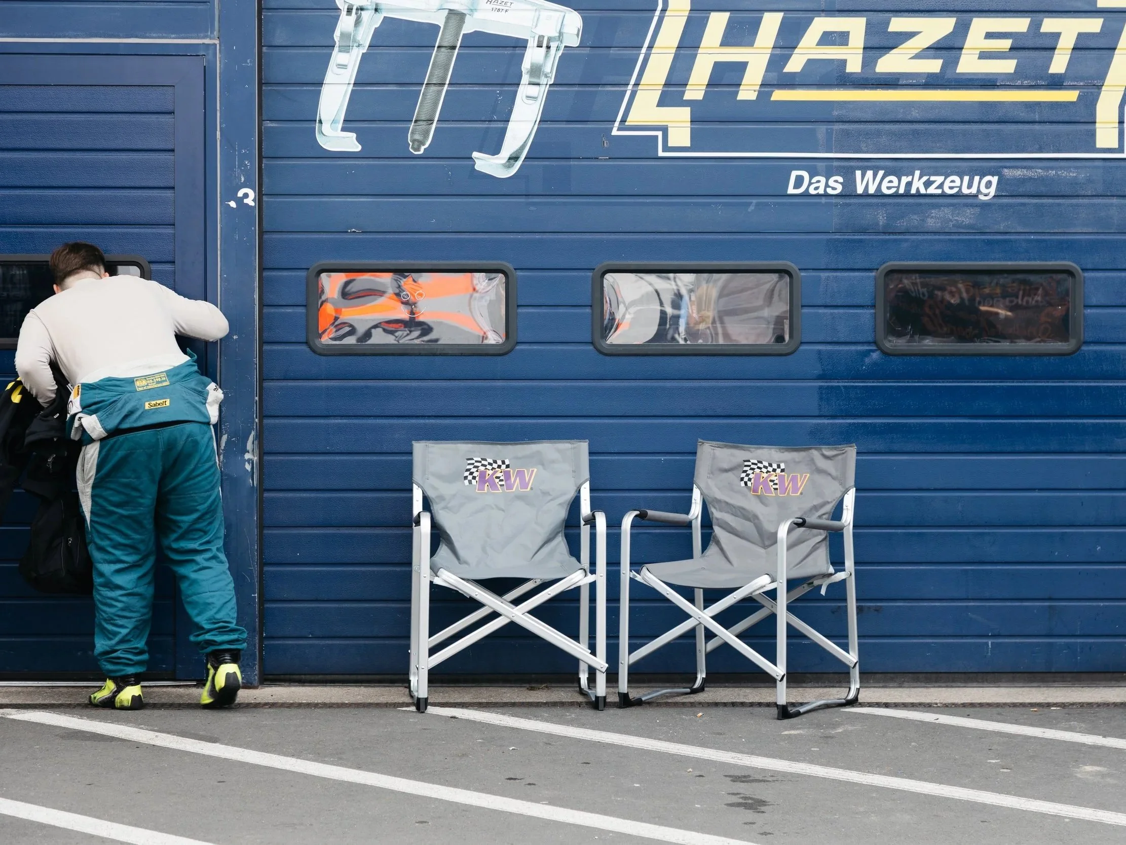 A blue garage door with four small windows and signage for 'Hazet' and 'Das Werkzeug'. Two folding chairs with 'KW' logos are in front of the door. A man in a white jacket and teal pants is standing beside a black bag, with his back to the camera.