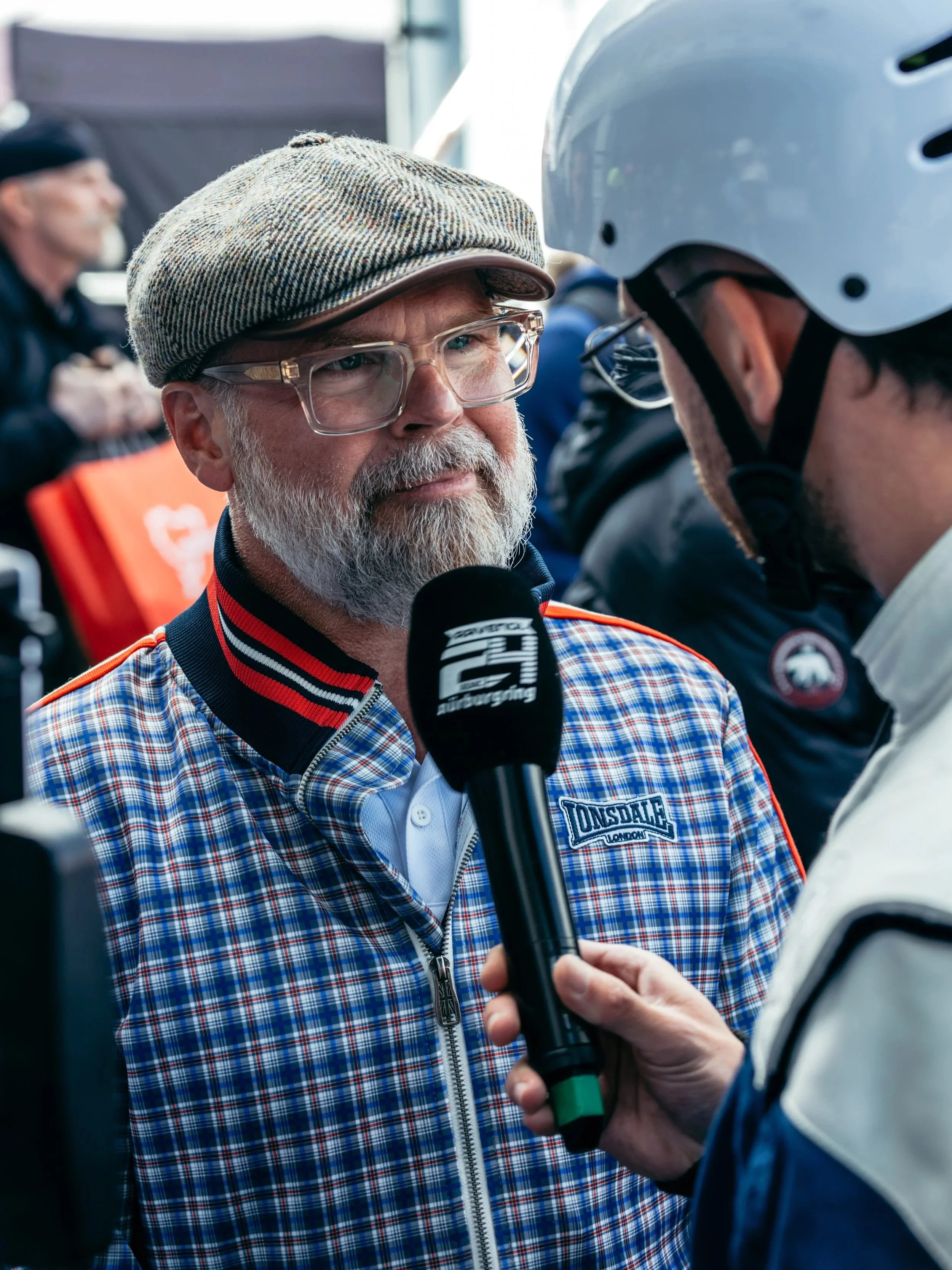 A man in a plaid jacket and glasses being interviewed with a microphone by a person wearing a white helmet.