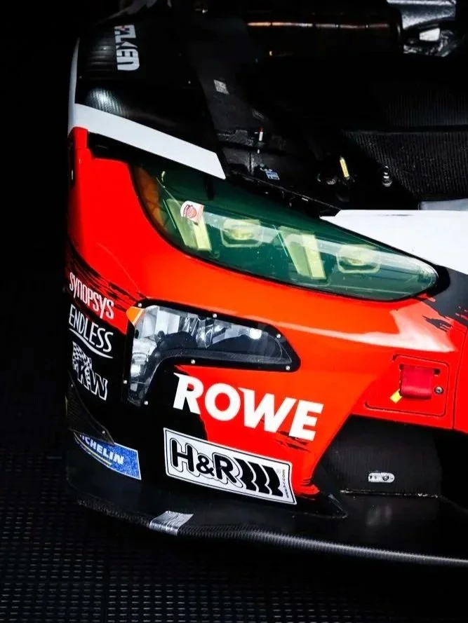 Close-up of the front section of a race car, featuring a red, black, and white color scheme with various sponsor logos including ROWE, H&R, Michelin, and Endless.