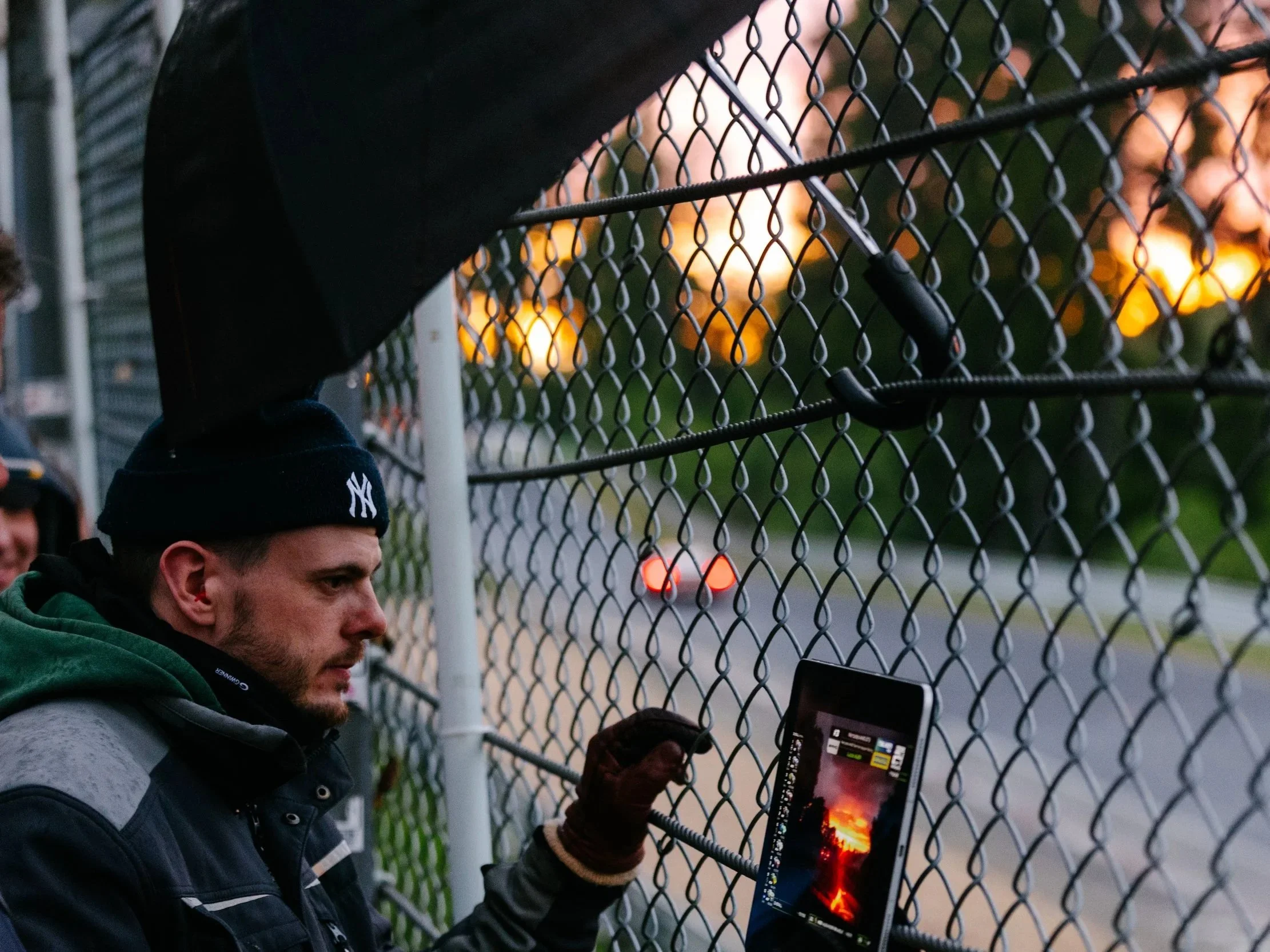 A young man wearing a dark jacket and a black beanie with a white NY logo, standing next to a chain-link fence at sunset. He is holding a tablet that displays a fiery landscape with volcanic activity.