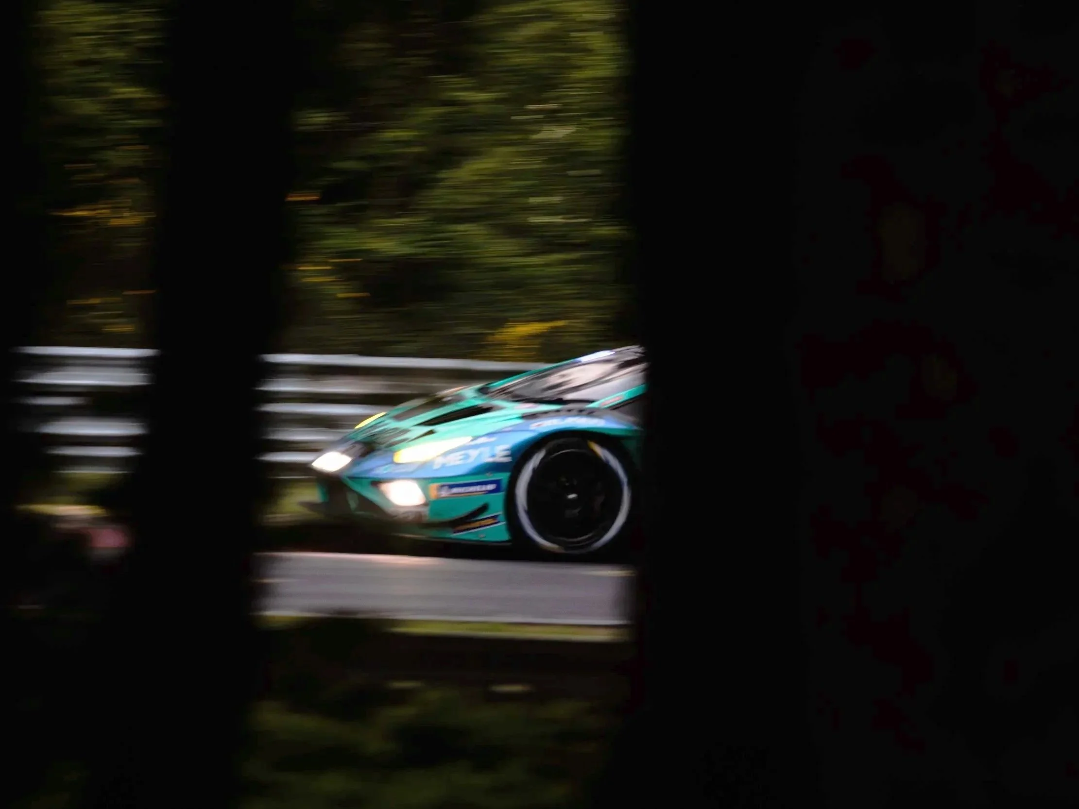 A race car speeding along a road, seen through a narrow gap between dark trees.
