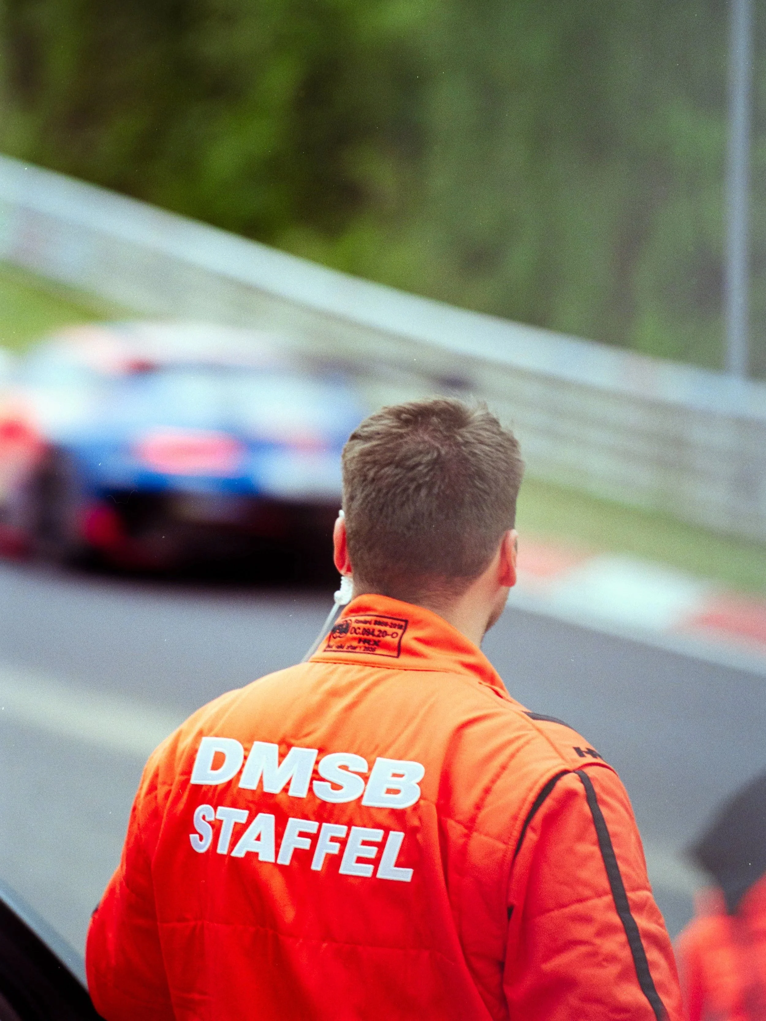 Race car driver in orange racing suit with 'DMSB STAFFEL' printed on the back, standing on a racetrack with racing cars blurred in the background.