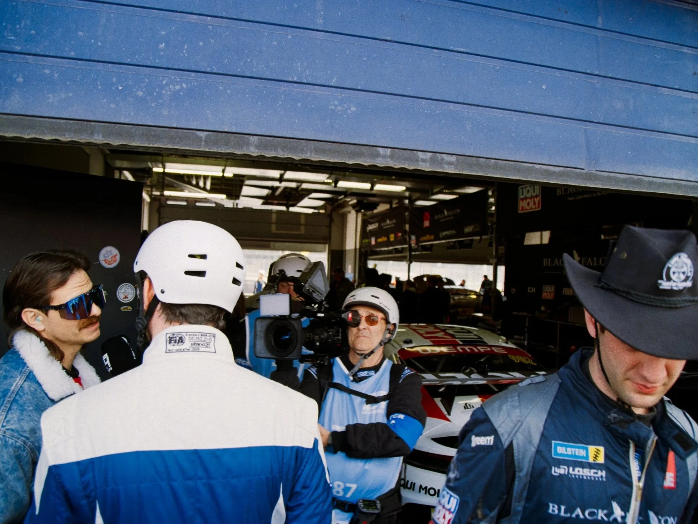 A group of people, including racers and team members, gathered at a race event, with some wearing helmets and racing gear, and an outdoor racing garage area in the background.