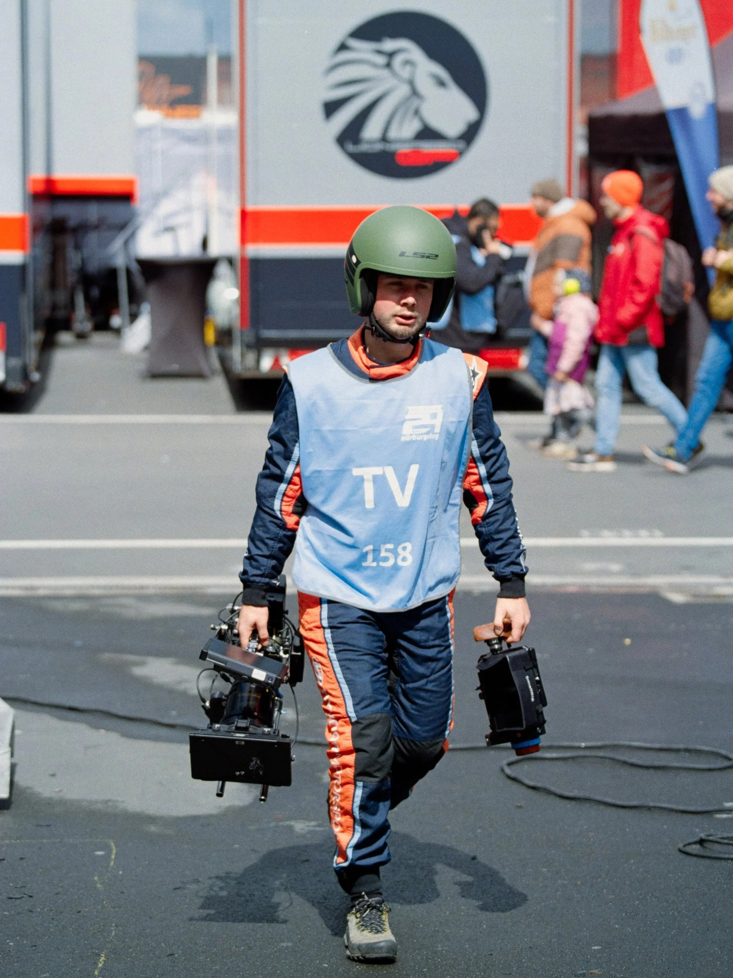 A man in racing gear with a green helmet walking with a drone in each hand at a racing event. Several people are in the background near a trailer with a lion logo.