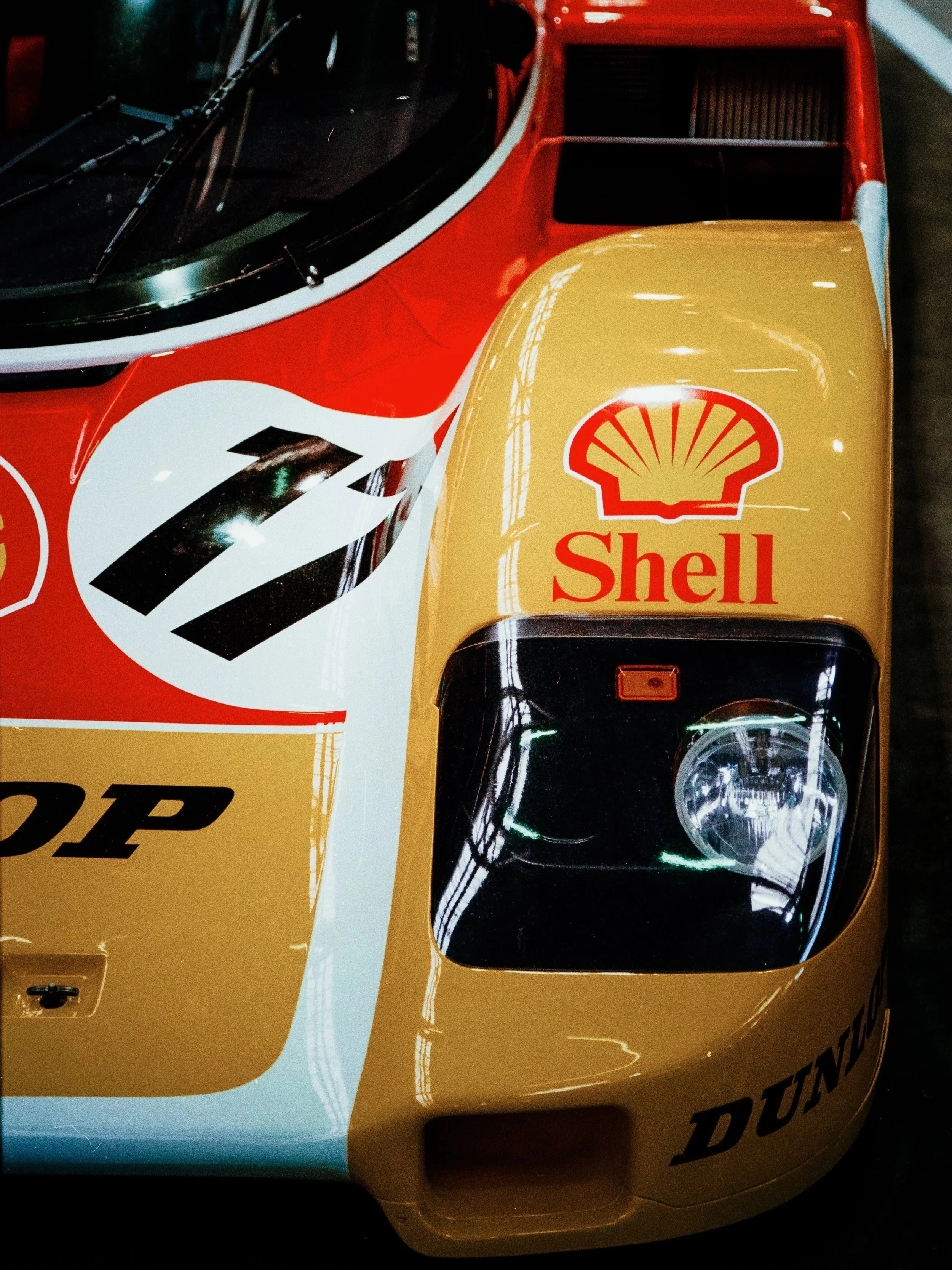 Close-up photo of the front section of a vintage race car with a yellow and red Shell logo on the hood, clear headlight, and aerodynamic design, taken from an overhead angle.