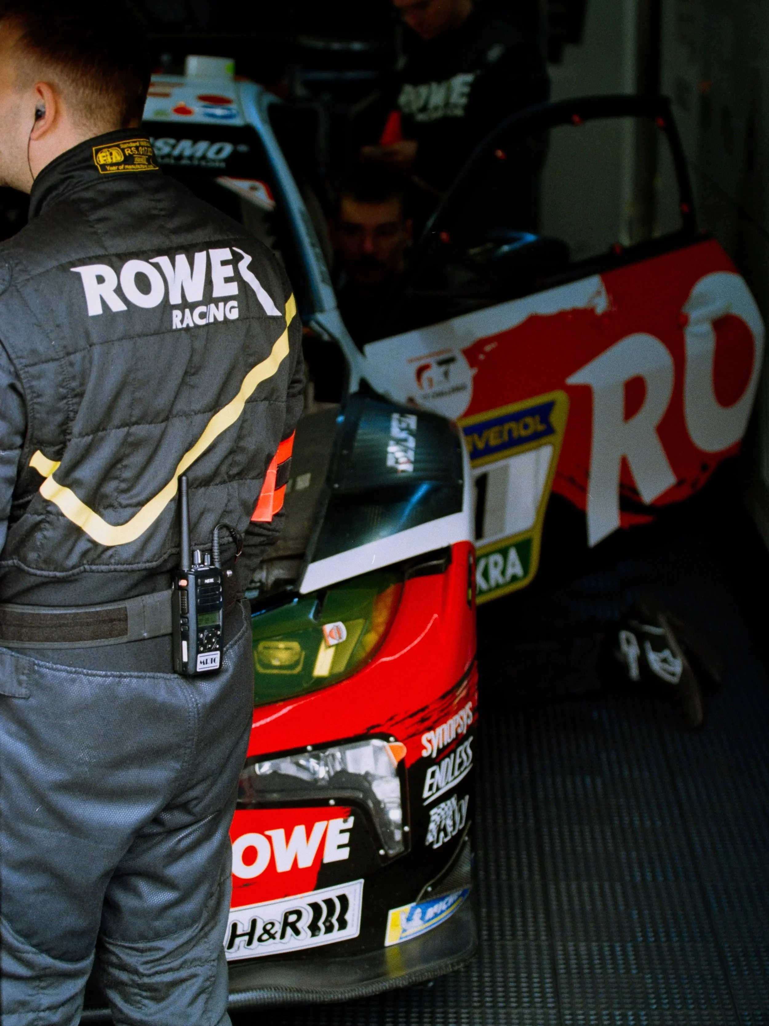 Pit crew members working on a race car in a garage, with one person in the foreground wearing a black racing suit with 'ROWES RACING' written on the back, and others in the background.