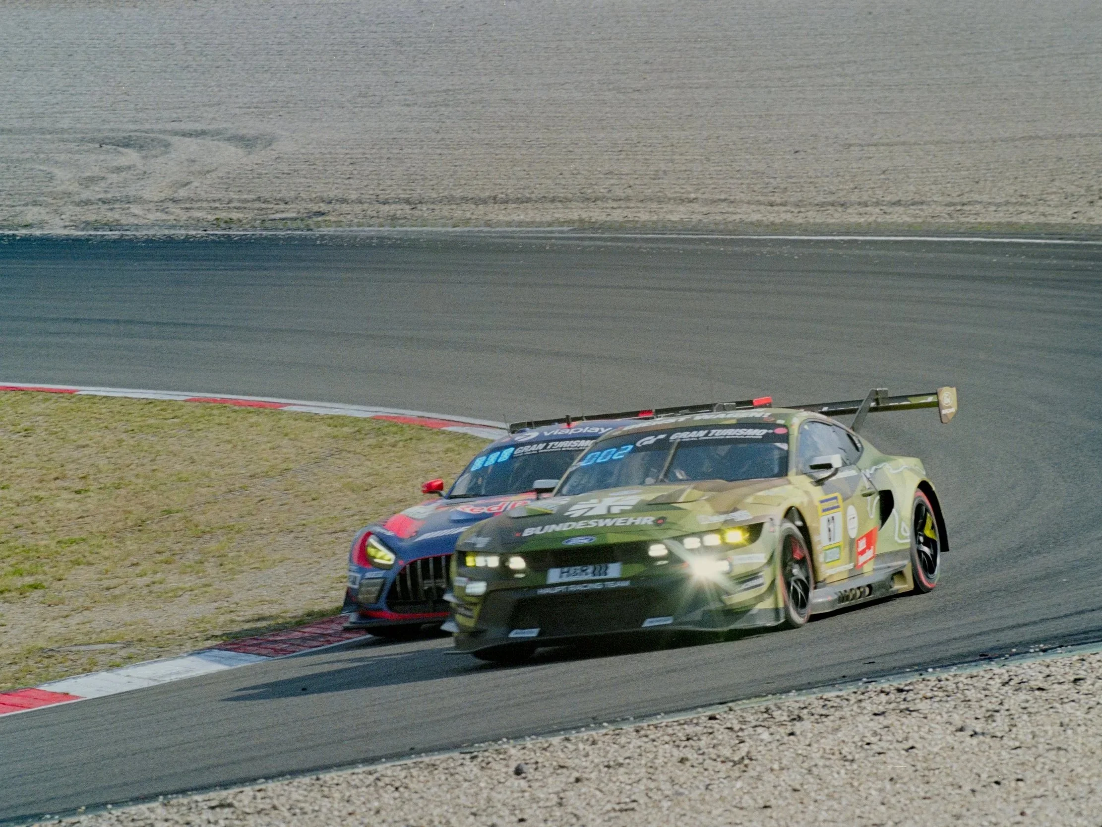 Two race cars on a racing track, with one orange and black car leading and a green car closely following, as they go around a curve on the track.