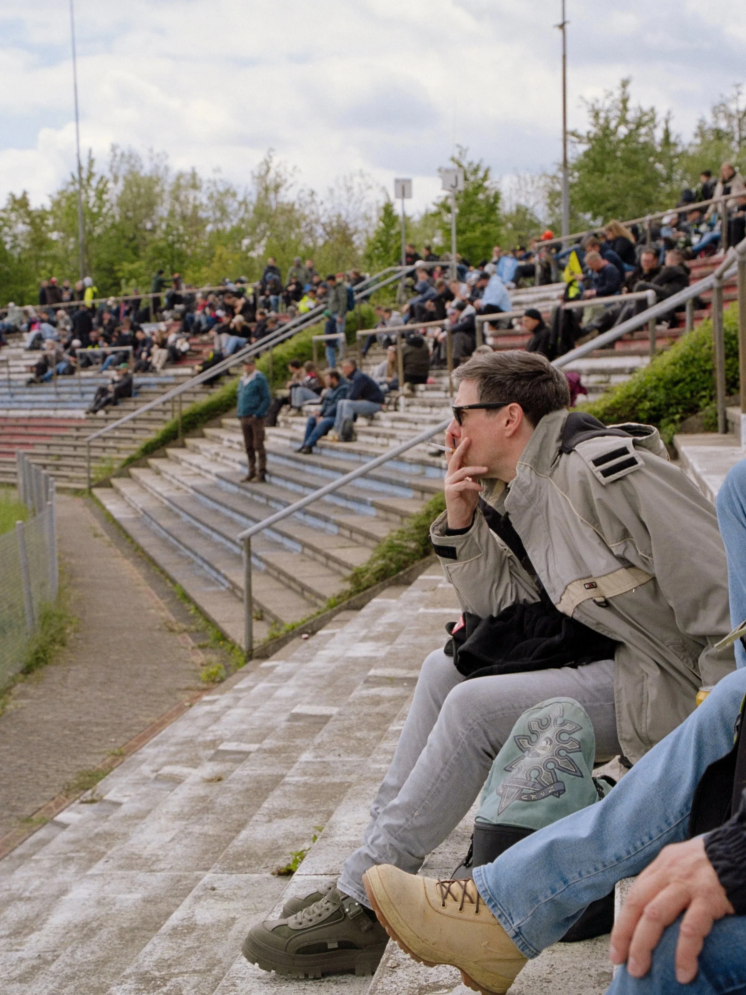 People sitting on bleachers outdoors on a cloudy day, some with backpacks, with trees and sky in the background.