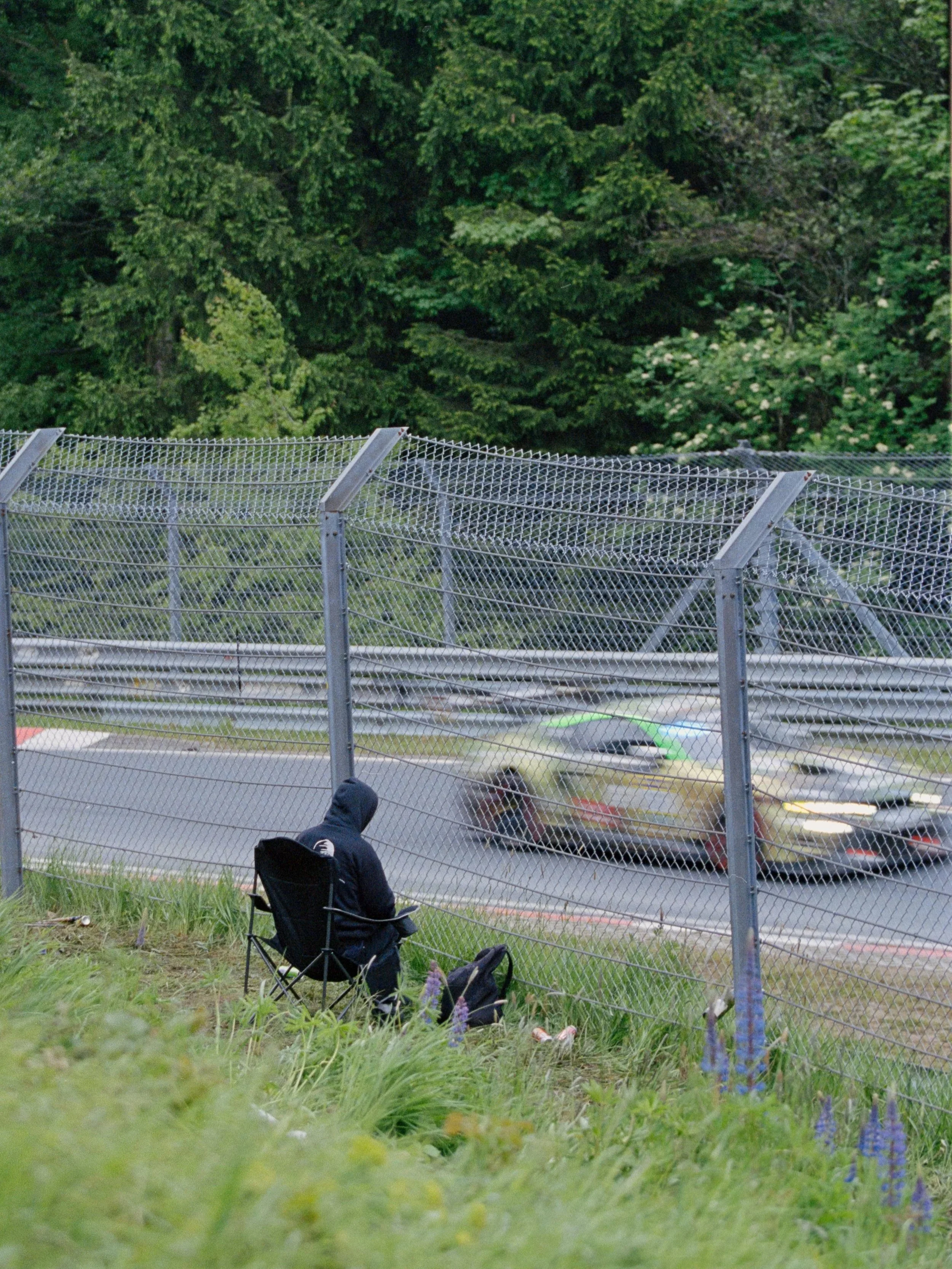 A person sitting on a folding chair by a chain-link fence, watching cars pass by on a road with greenery and trees in the background.