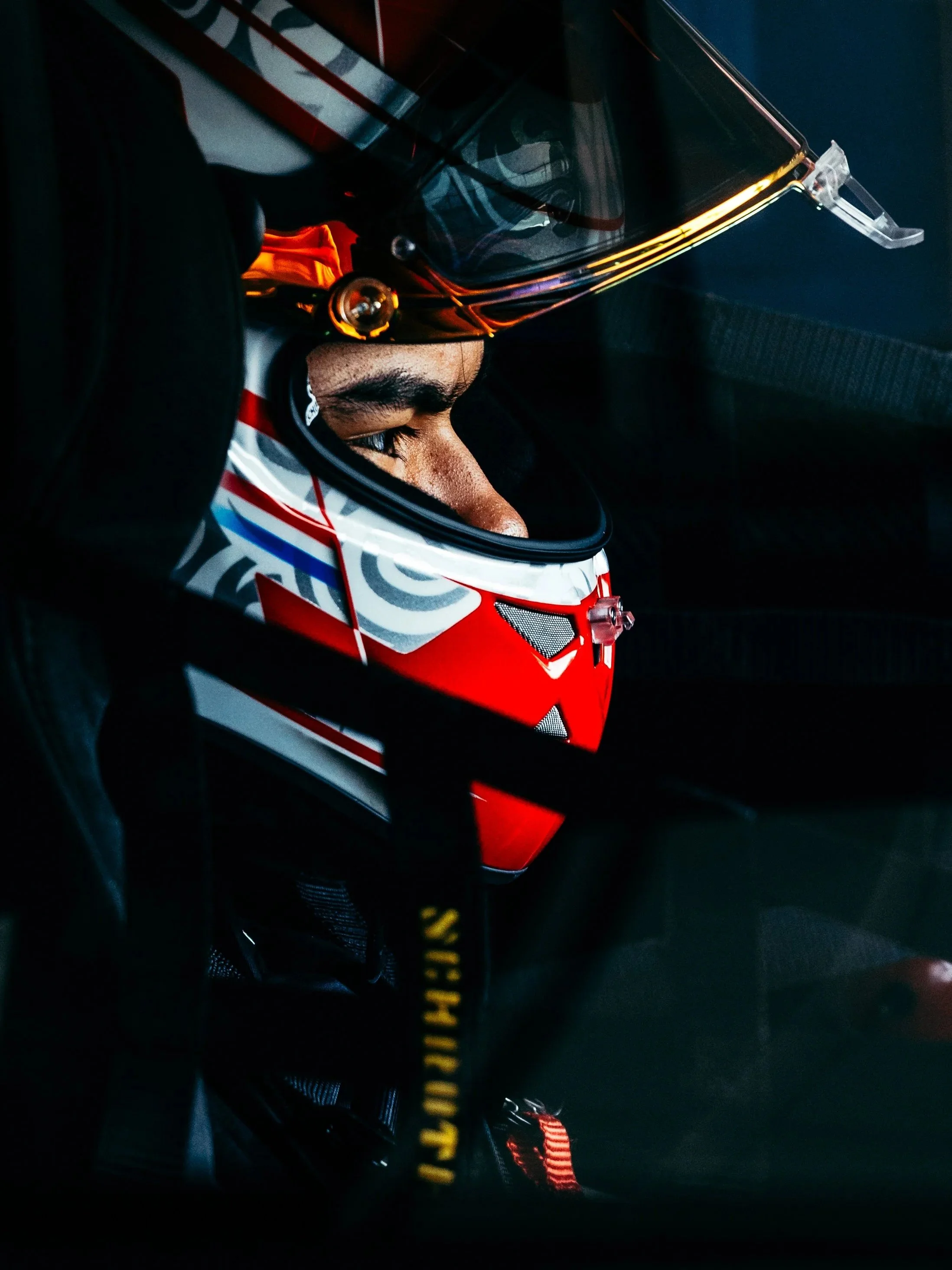 Close-up of a race car driver in a helmet with a reflective visor, inside a dark vehicle with a red and white design.