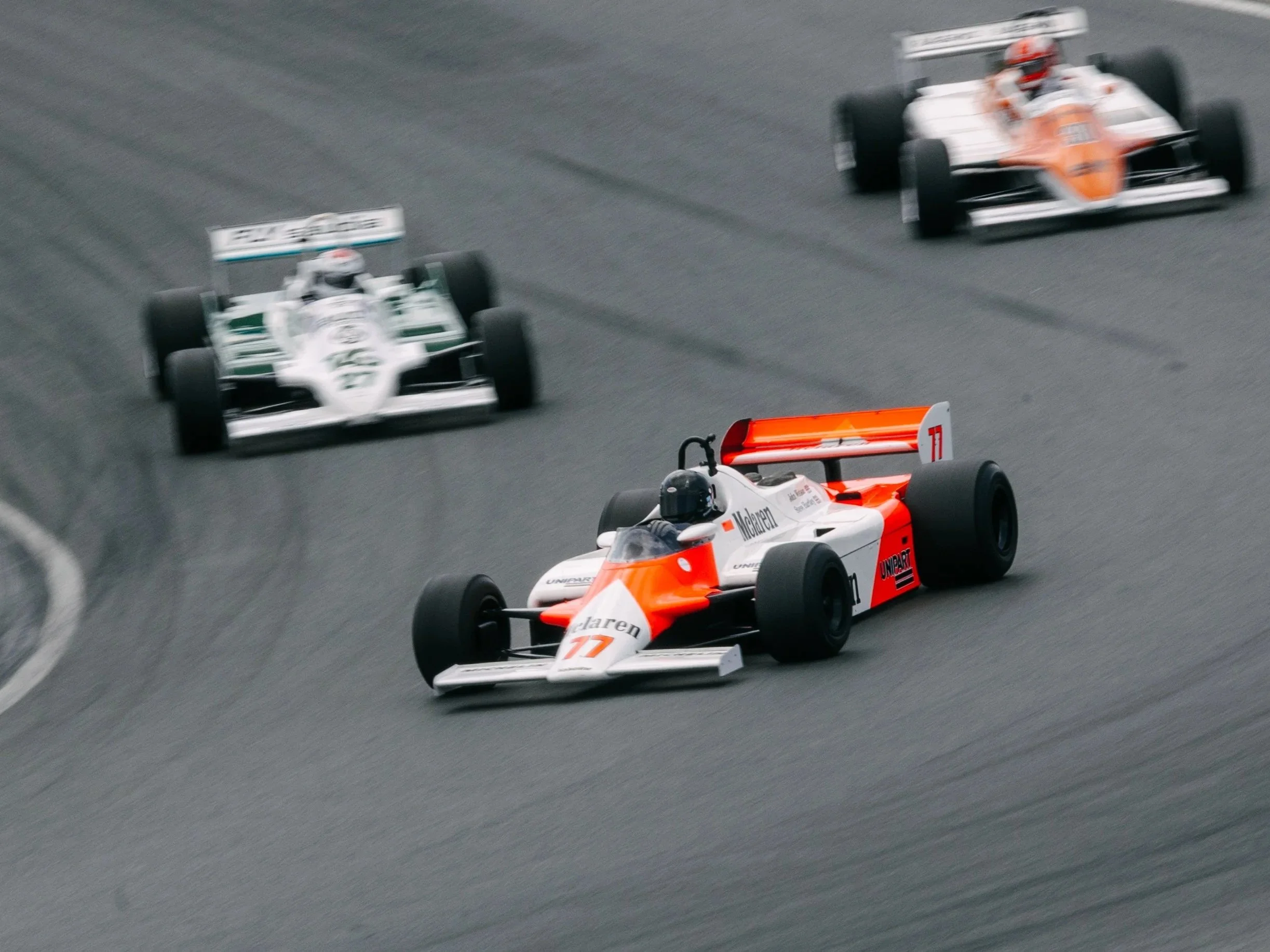 Three Formula 1 race cars on a racetrack, with the front car being McLaren and the two cars behind it appearing to be part of a different team. The cars are in motion on a gray asphalt track.