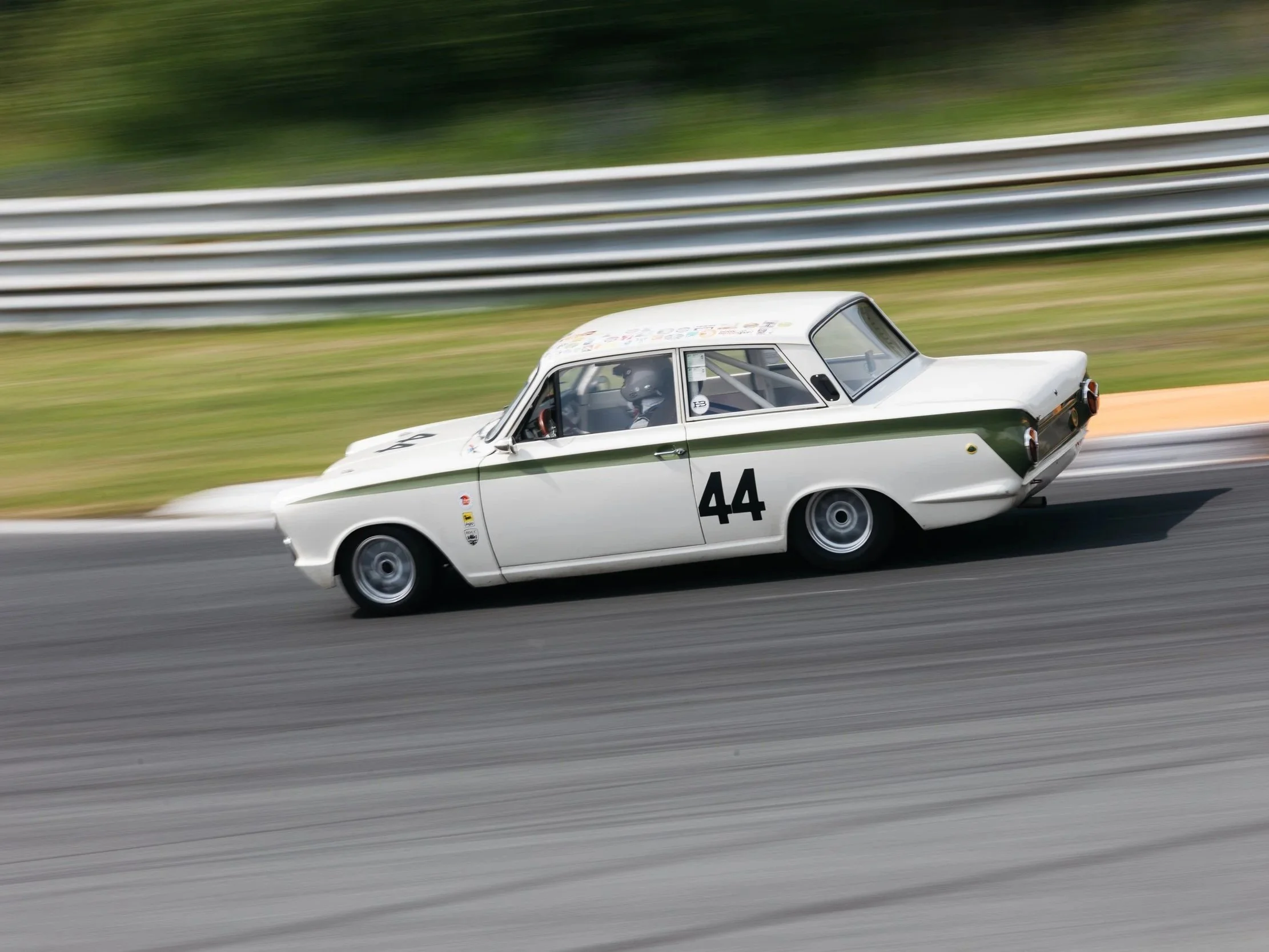 A white vintage race car with the number 44 on its side speeds along a racetrack, with blurred grass and metal guardrails in the background.