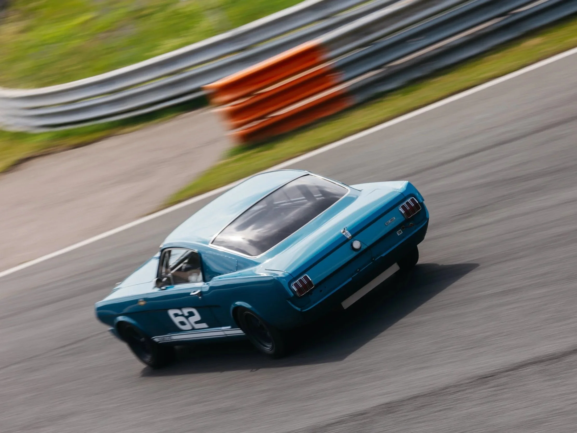 A blue vintage car with racing decals, number 62, speeding on a race track with a blurred background and fencing.