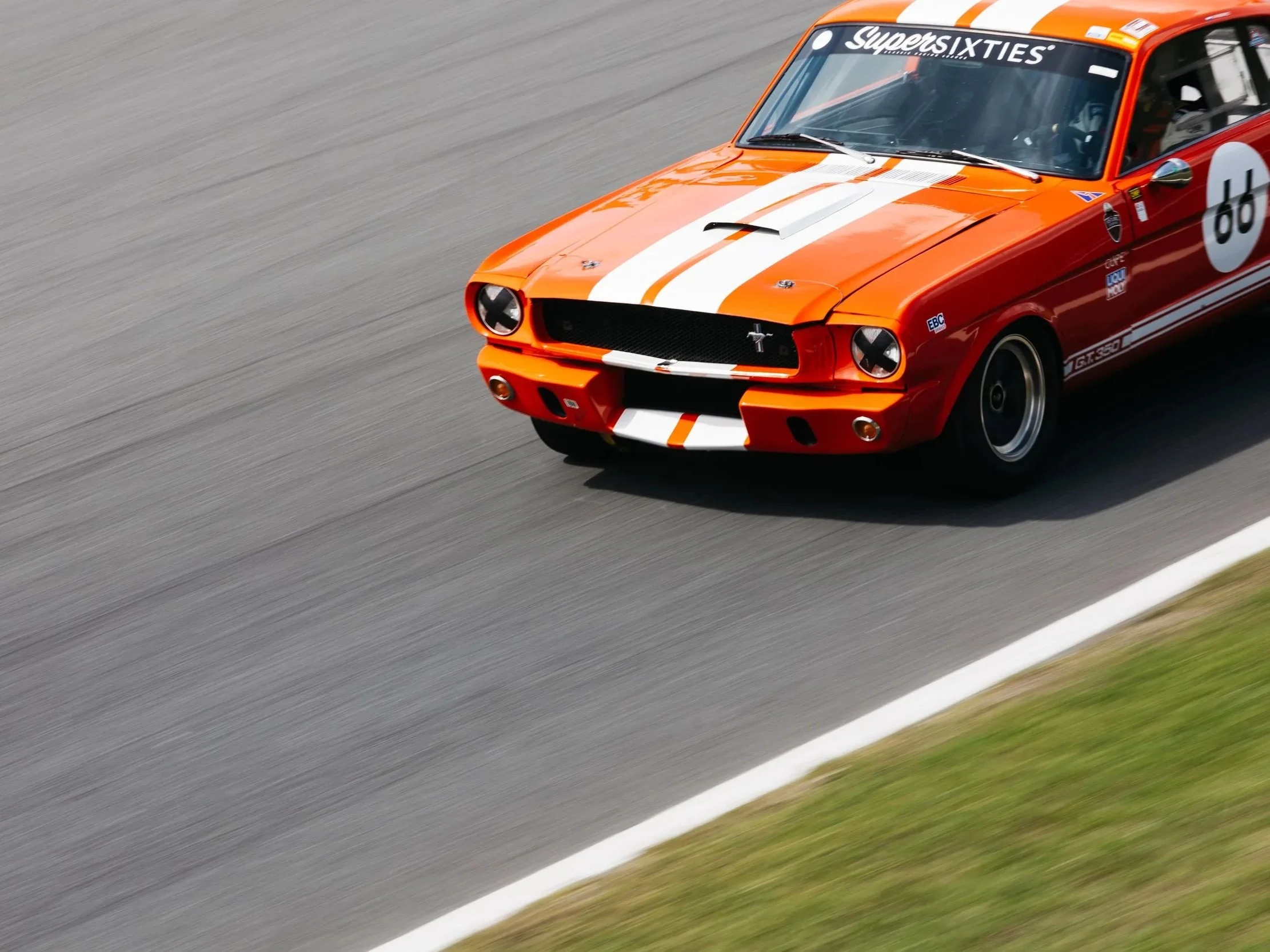 A bright orange vintage race car with white racing stripes driving on a race track.