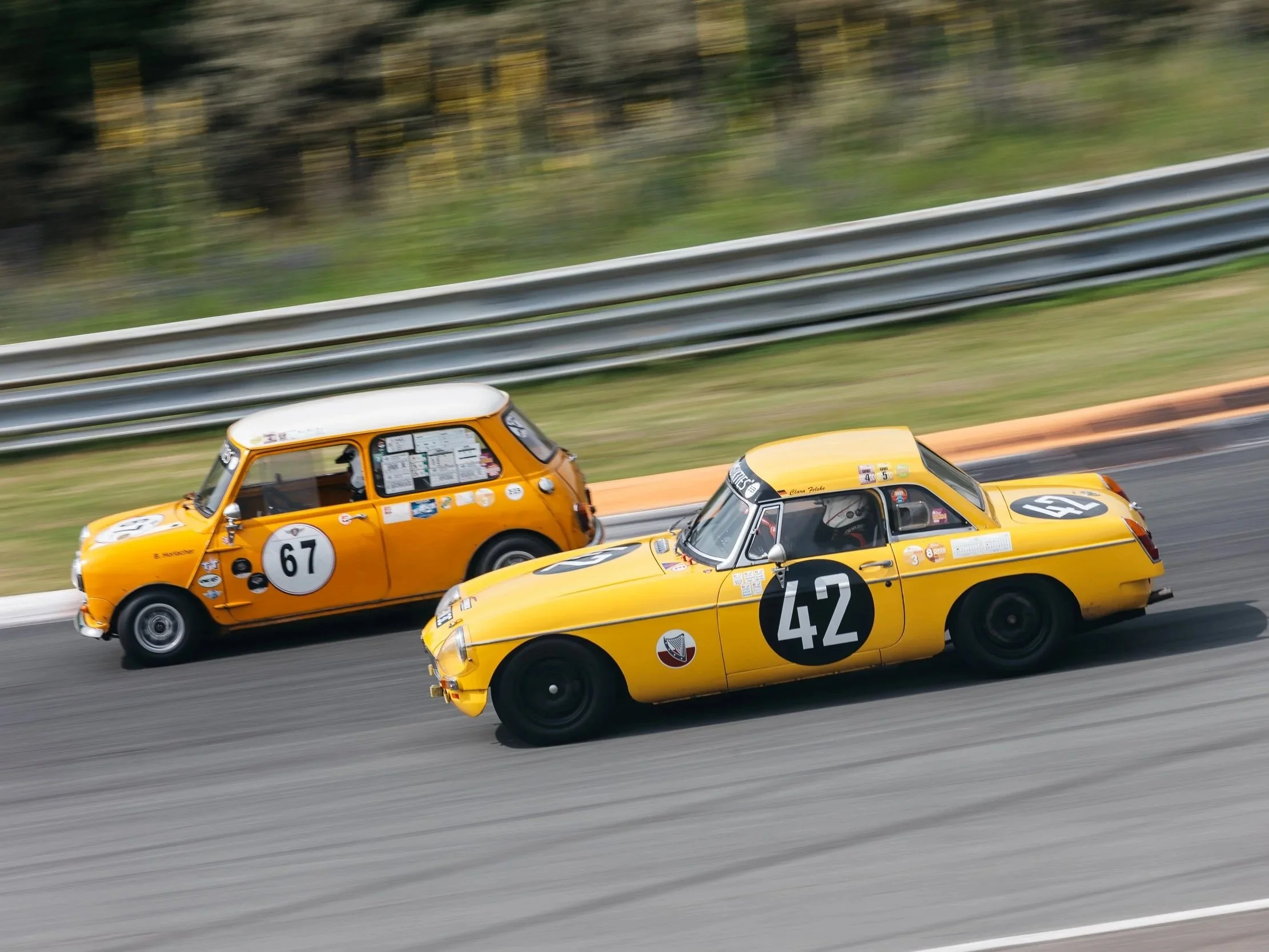 Two yellow race cars with racing numbers, #67 and #42, driving on a track with a metal guardrail and trees in the background.