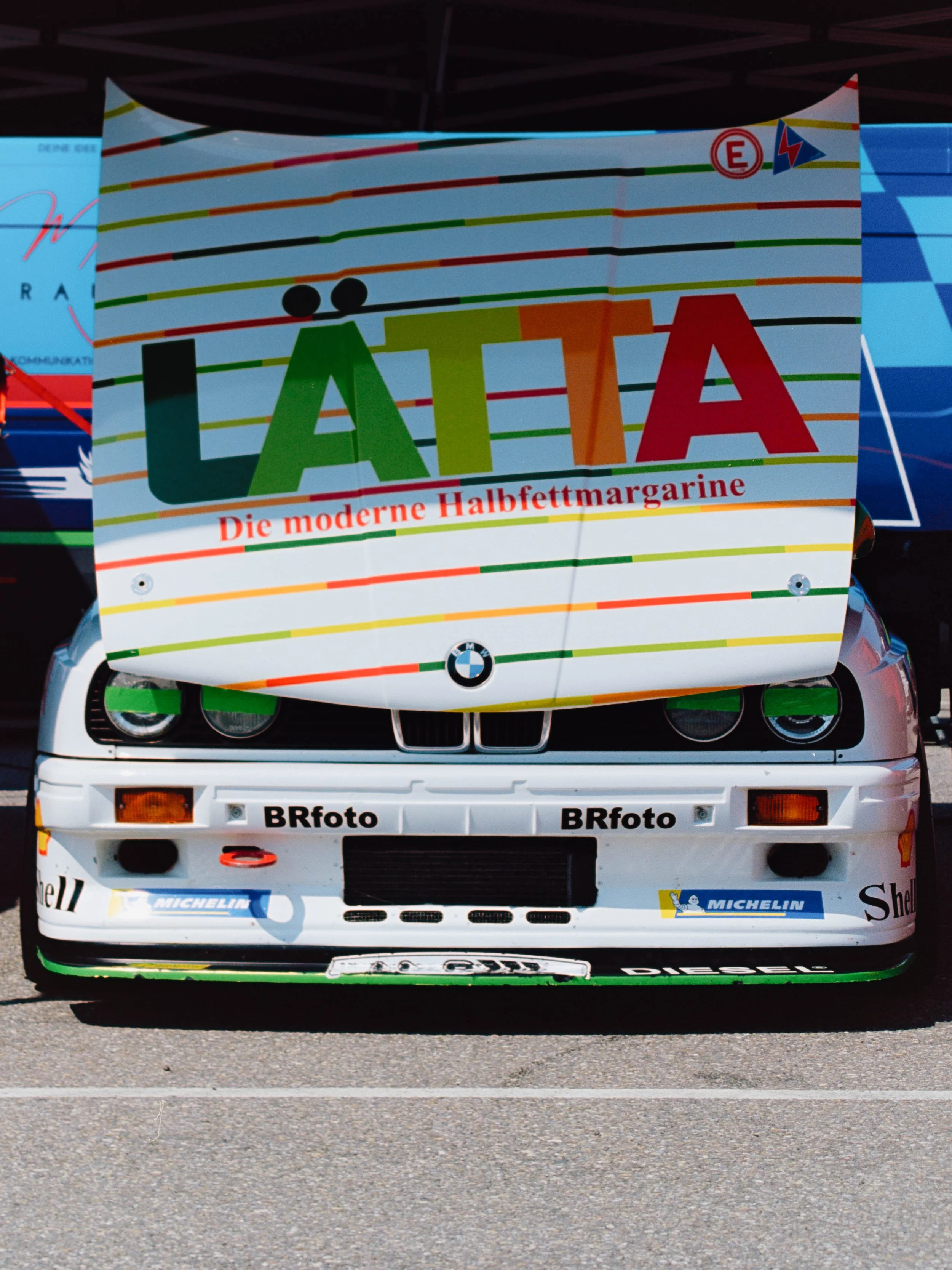 Front view of a white race car with a large colorful advertising banner on the hood reading 'Lätta,' with the subtitle 'Die moderne Halbfettmargarine,' and various logos including BMW, Michelin, Shell, and Dsiegel.