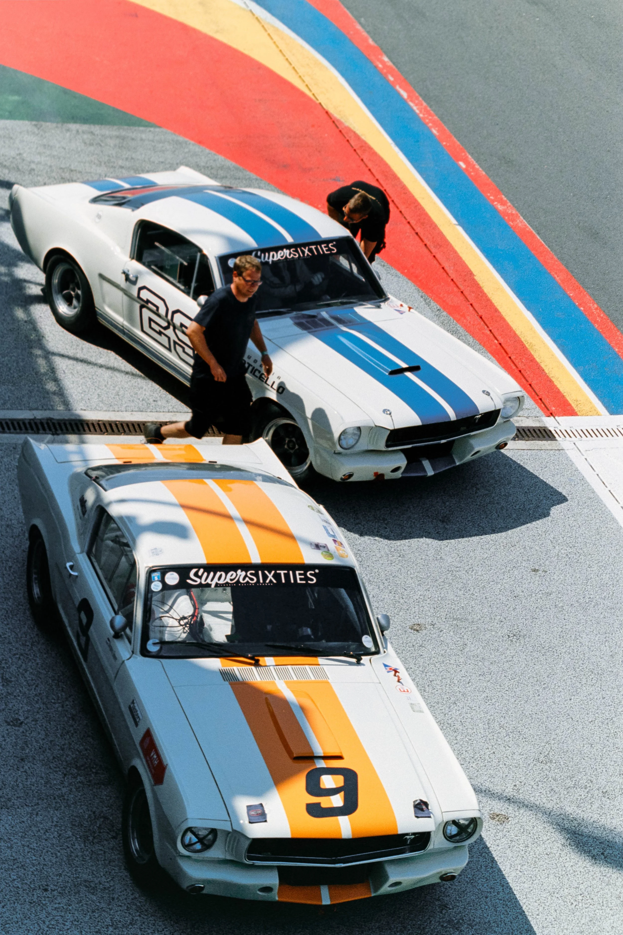 Two vintage white race cars with orange and blue stripes parked on a race track, with two people attending to one of the cars.