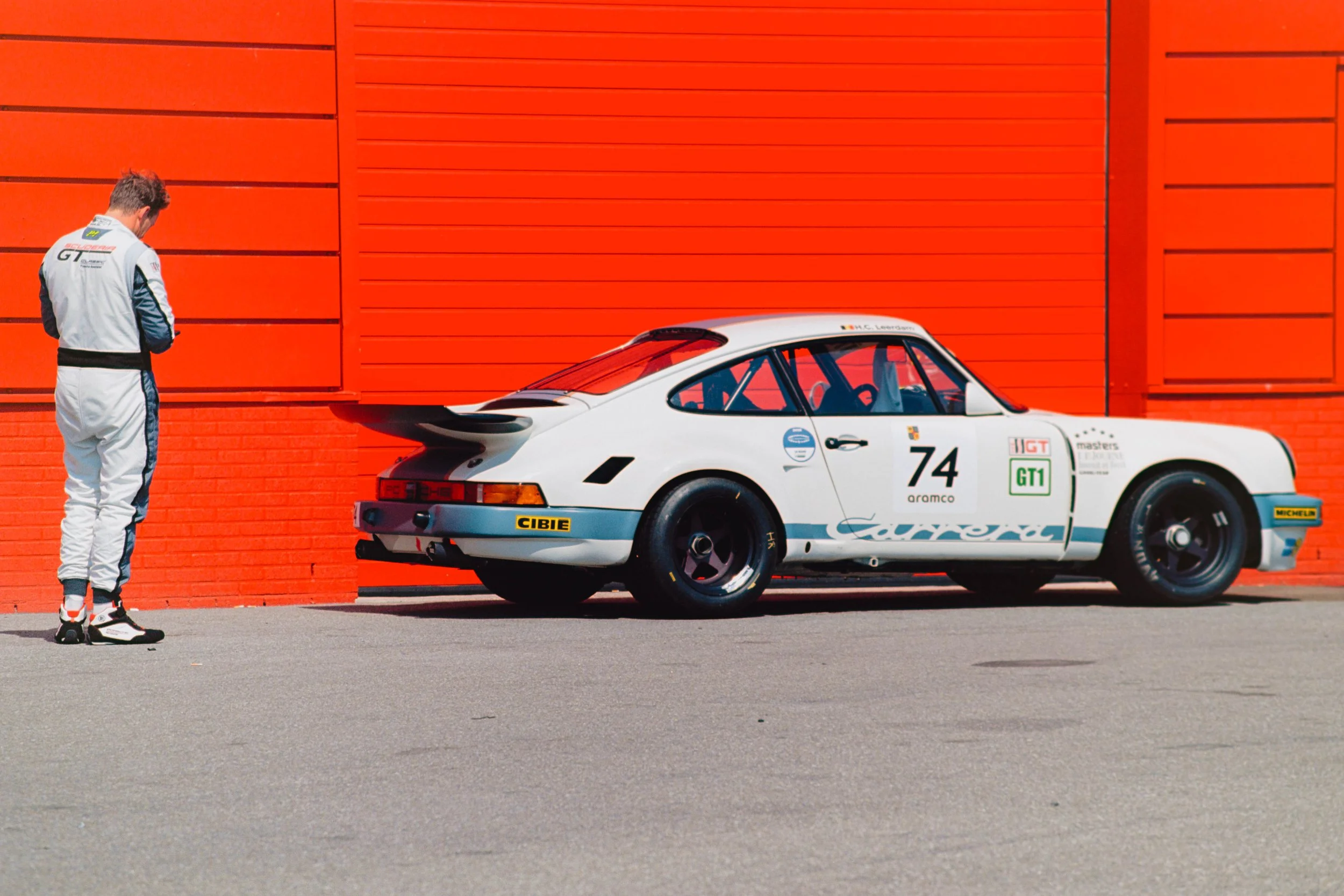 A man dressed in racing gear standing near a classic white Porsche race car with racing decals, parked in front of a bright orange building.