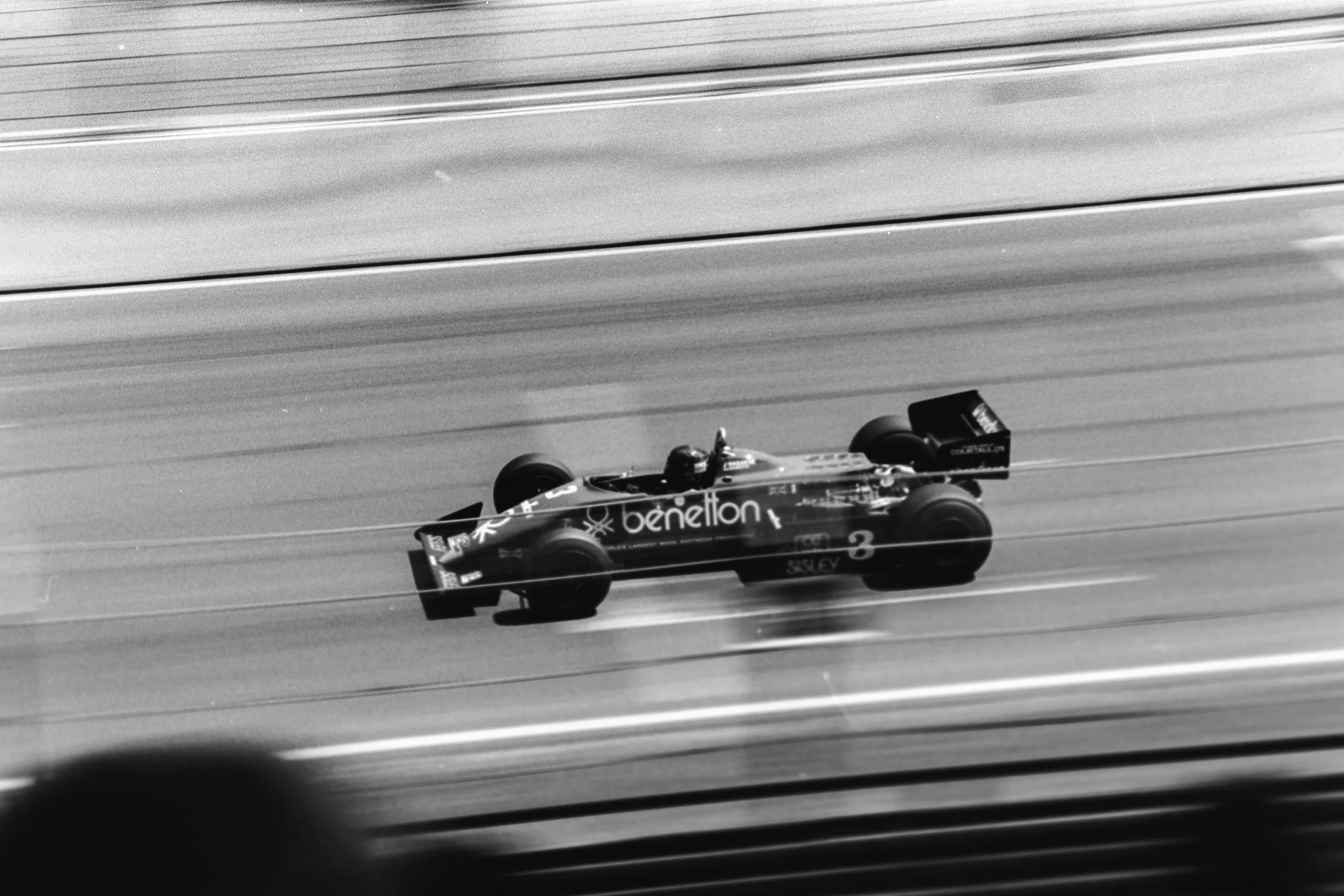 Black and white photo of a race car speeding on a track, viewed from a distant perspective, with the background showing several horizontal lines likely representing the race track surface.